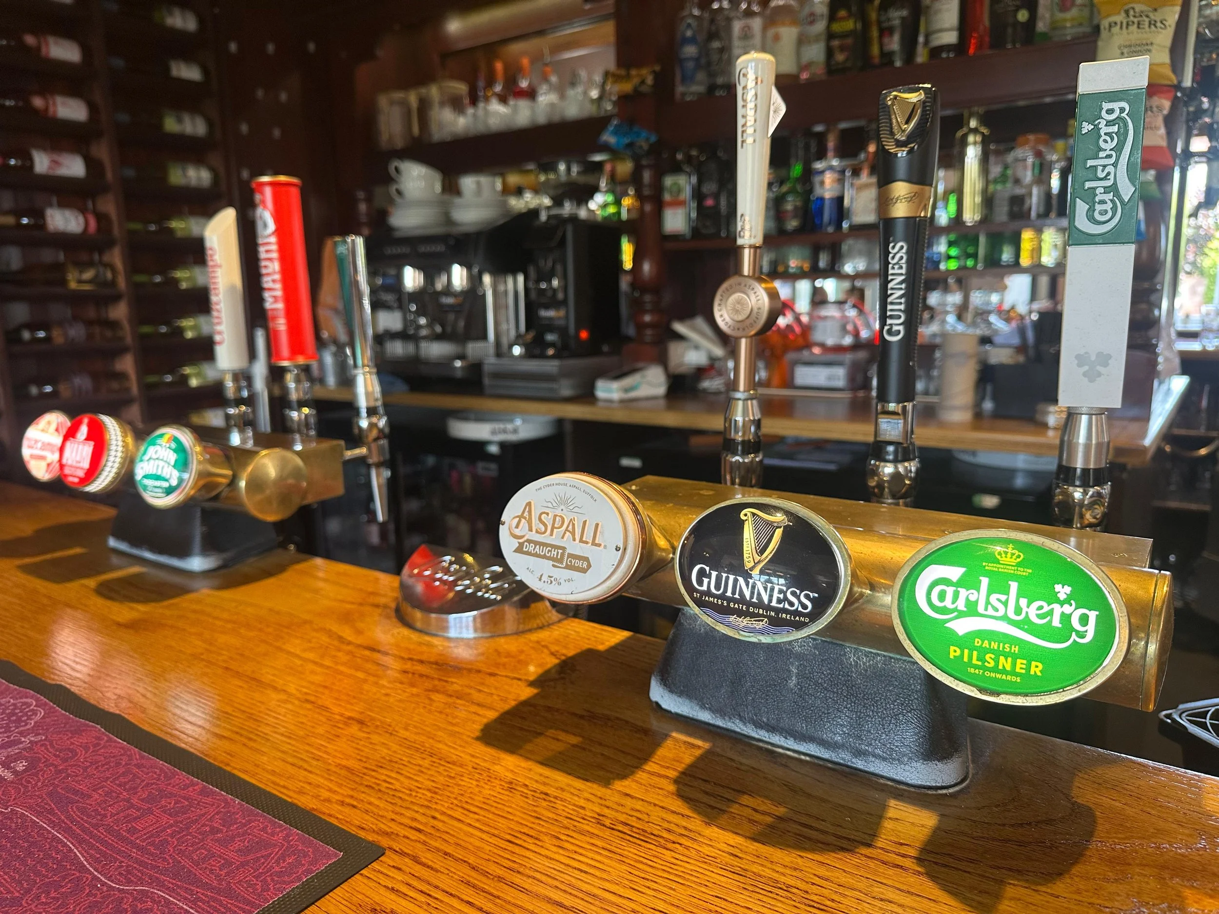 Bar with beer tap handles featuring brands such as Aspall, Guinness, and Carlsberg, on a wooden counter in a pub or bar setting.