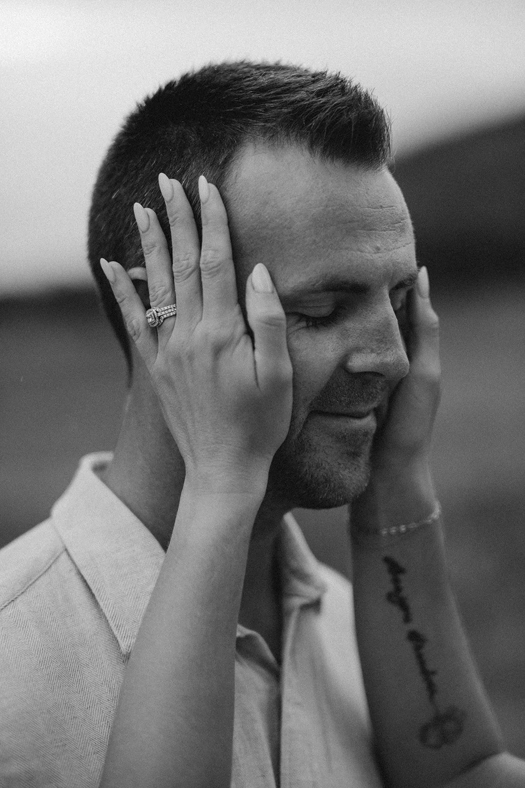A black-and-white photo of a man with short hair, eyes closed, holding his face with two hands. A woman’s hands are on his face, one with a ring on the finger. The woman’s arms are visible, with a tattoo on one forearm. The man appears to be in a peaceful or emotional moment.