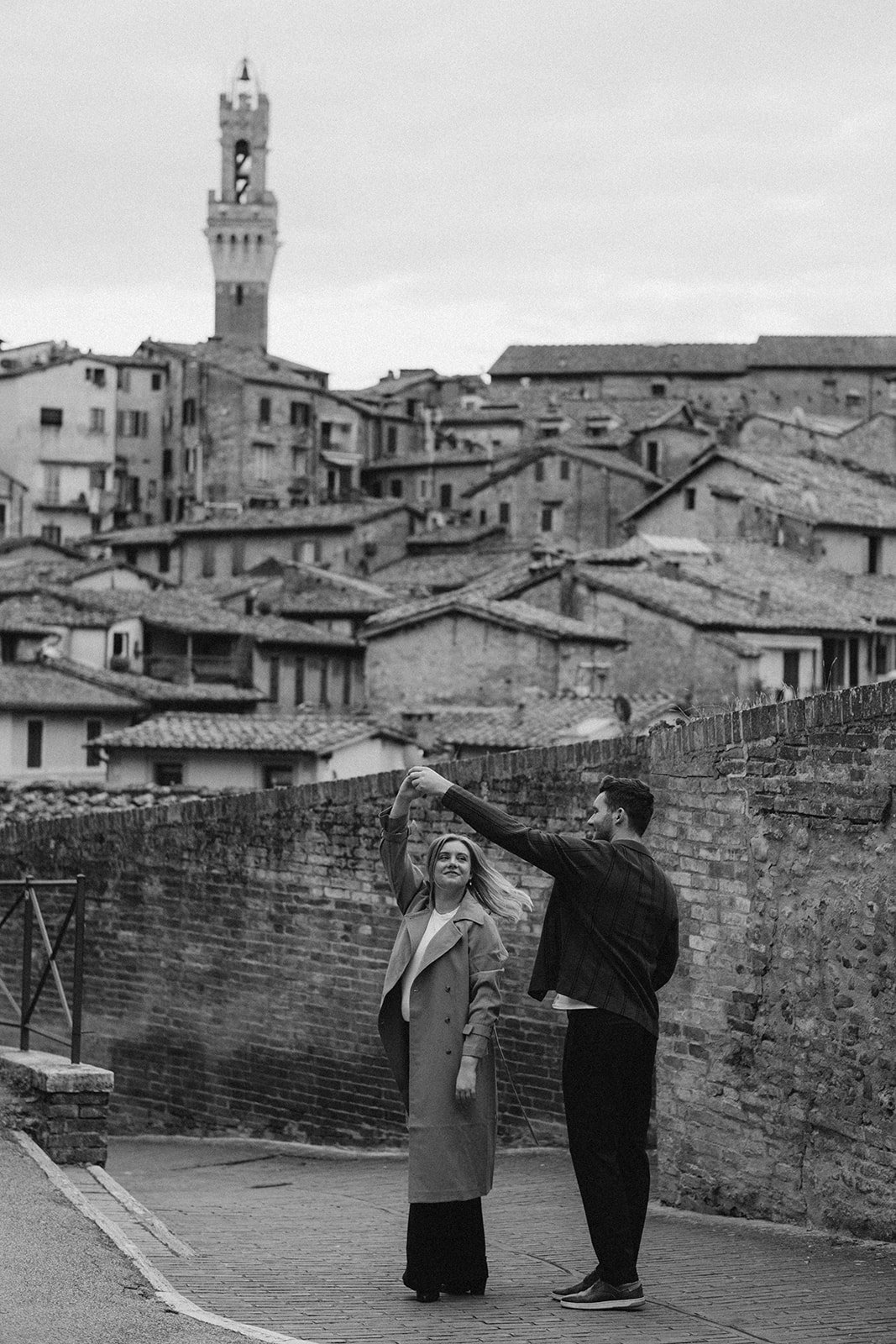 A couple dancing on a cobblestone street against the backdrop of Siena's old Italian townhouses and the clock tower of Piazza del Campo.