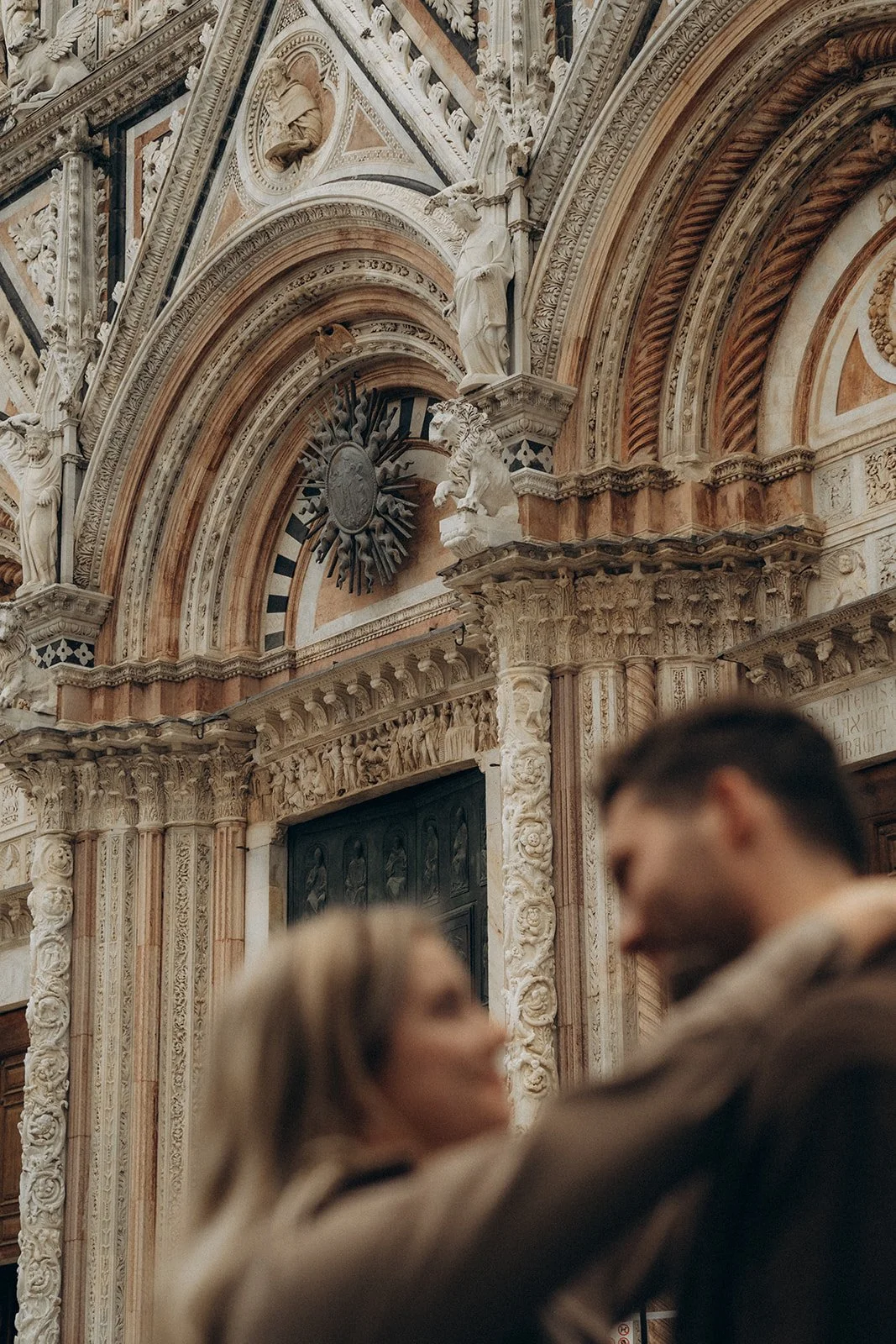 Blurred image of a man and woman dancing or embracing in front of ornate, classical-style architectural interior with intricate carvings, statues, and decorative elements of the Siena Cathedral.