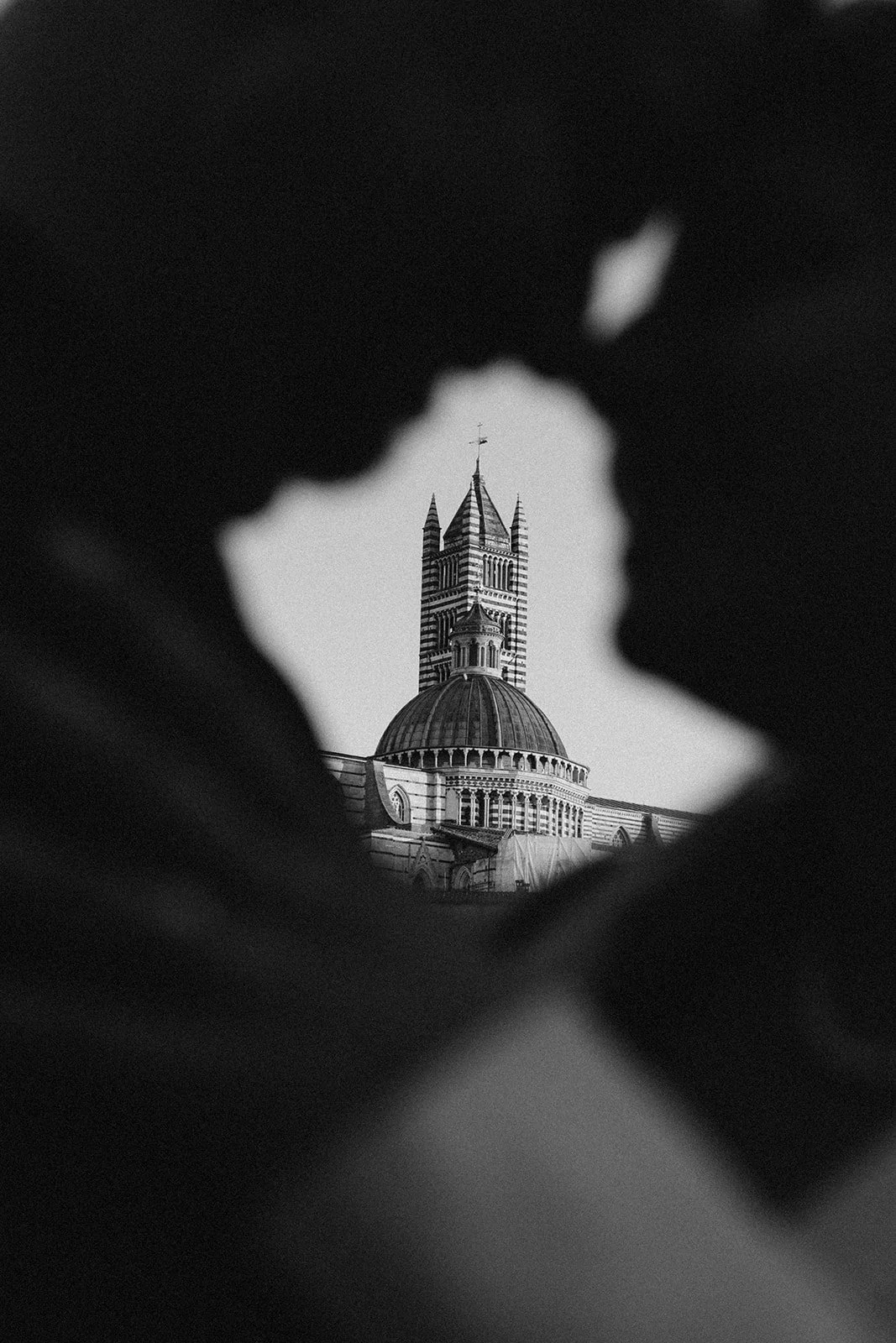 Black and white photo showing the Cathedral of Siena through an opening between two people.
