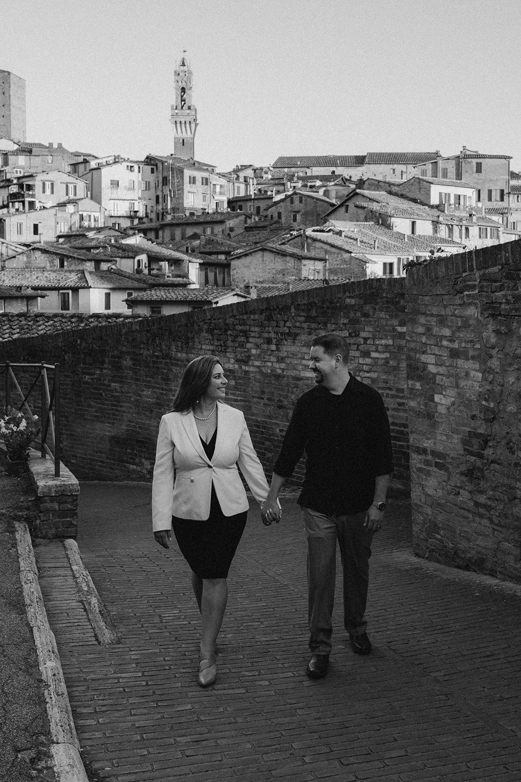 A woman and man holding hands and walking together on a brick pathway in Siena, with a hillside of tightly packed buildings and the Torre del Mangia tower in the background, in black and white.