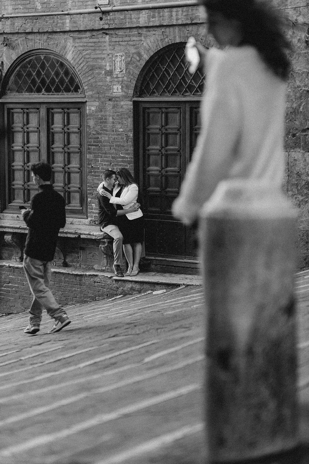 A black-and-white photo of a brick building with arched windows and closed wooden doors. In the foreground, a woman leans against the wall as she and a man share a kiss. Two other people, a man and a woman, are walking nearby.