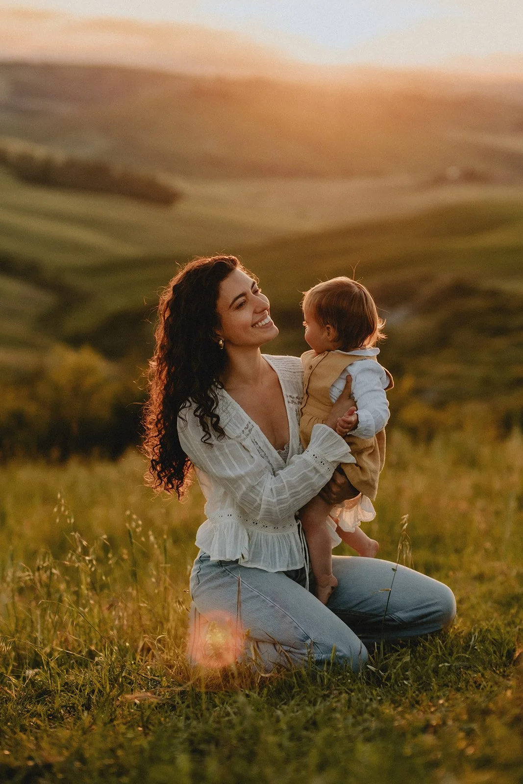 A woman kneeling in a grassy field, holding a young child in her arms during sunset, with rolling hills in the background.