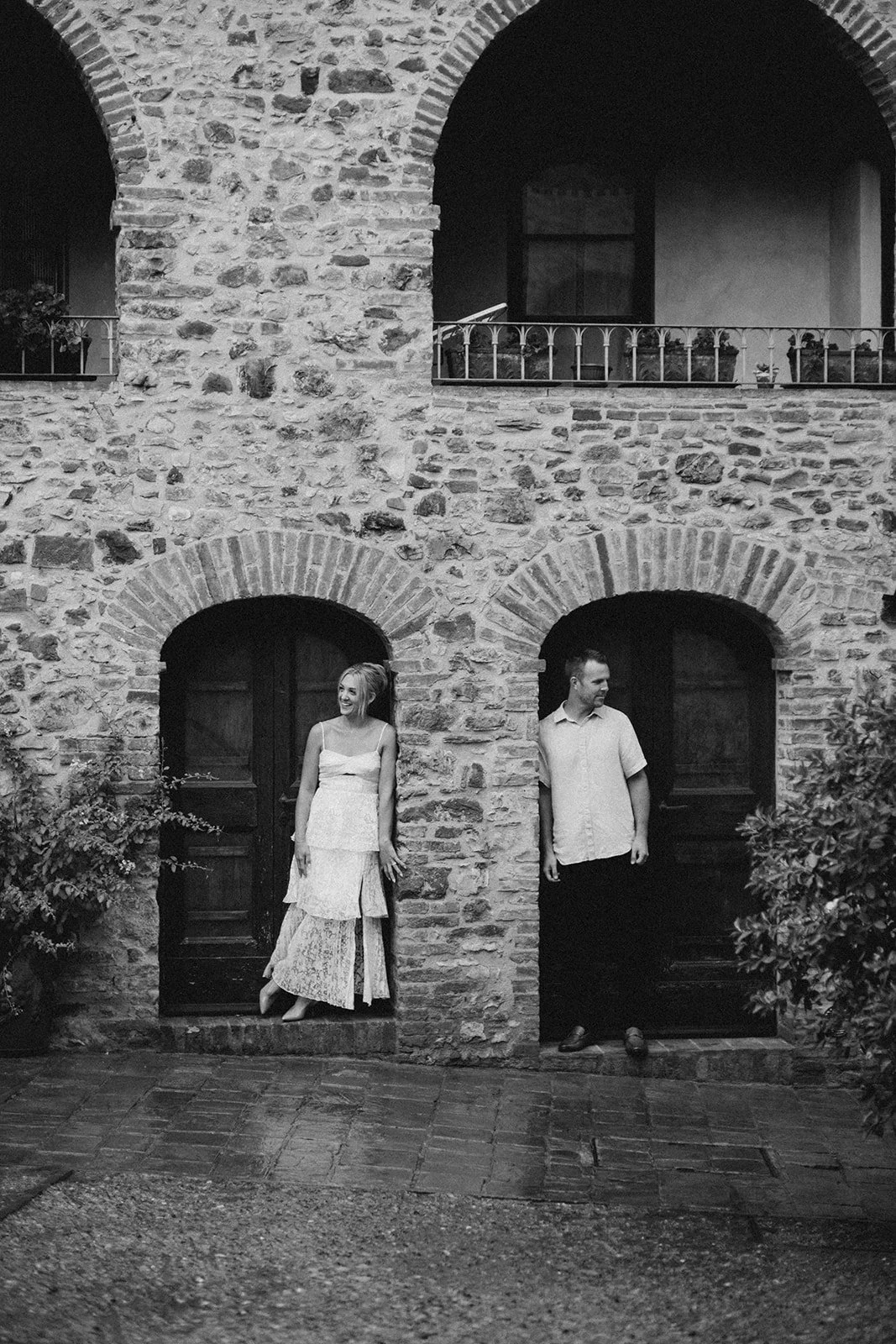 A black and white photo of a stone building with three arches, two on the ground level and one on the upper level. Two people, a woman and a man, are standing in the middle arches, smiling and looking at each other. The woman is on the left, wearing 