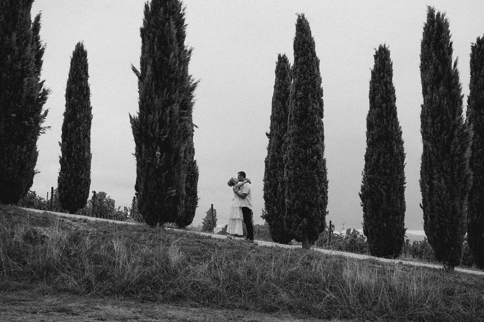 A couple embracing on a pathway surrounded by tall cypress trees, in black and white.