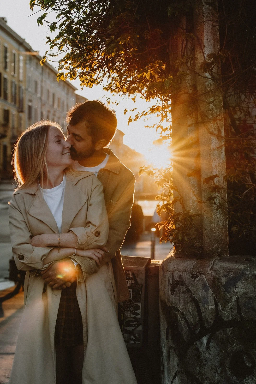 A couple embraces and kisses on a city street at sunset, with buildings and a stone wall with graffiti nearby.