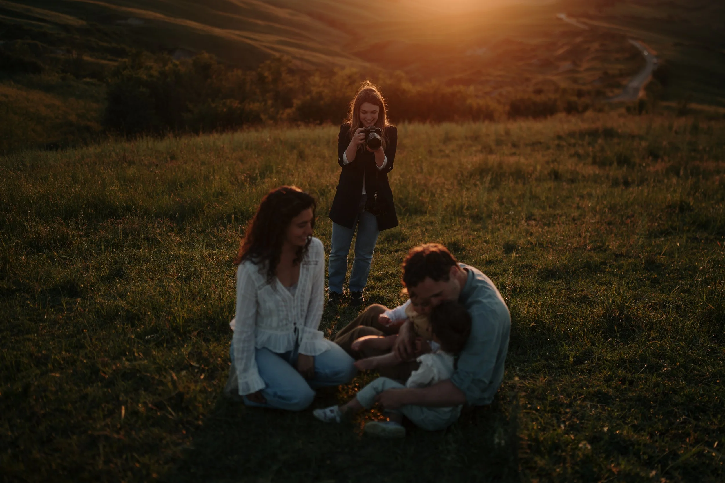 Family of four sitting and hugging in a grassy field during sunset, with a Siena Hilt Photography taking a photo in the background.