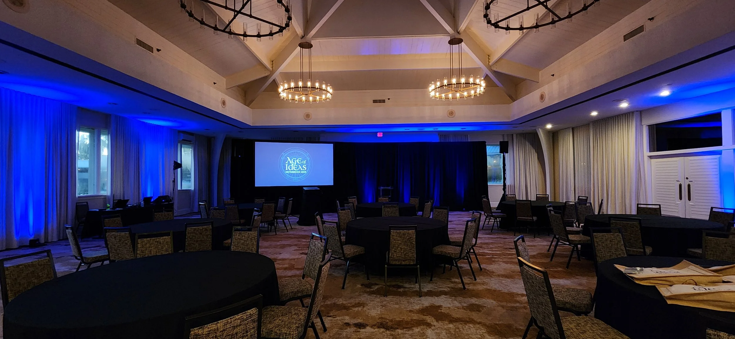 Conference room with round tables and chairs, a large screen displaying 'Age of Ideas' logo, and blue lighting accents.