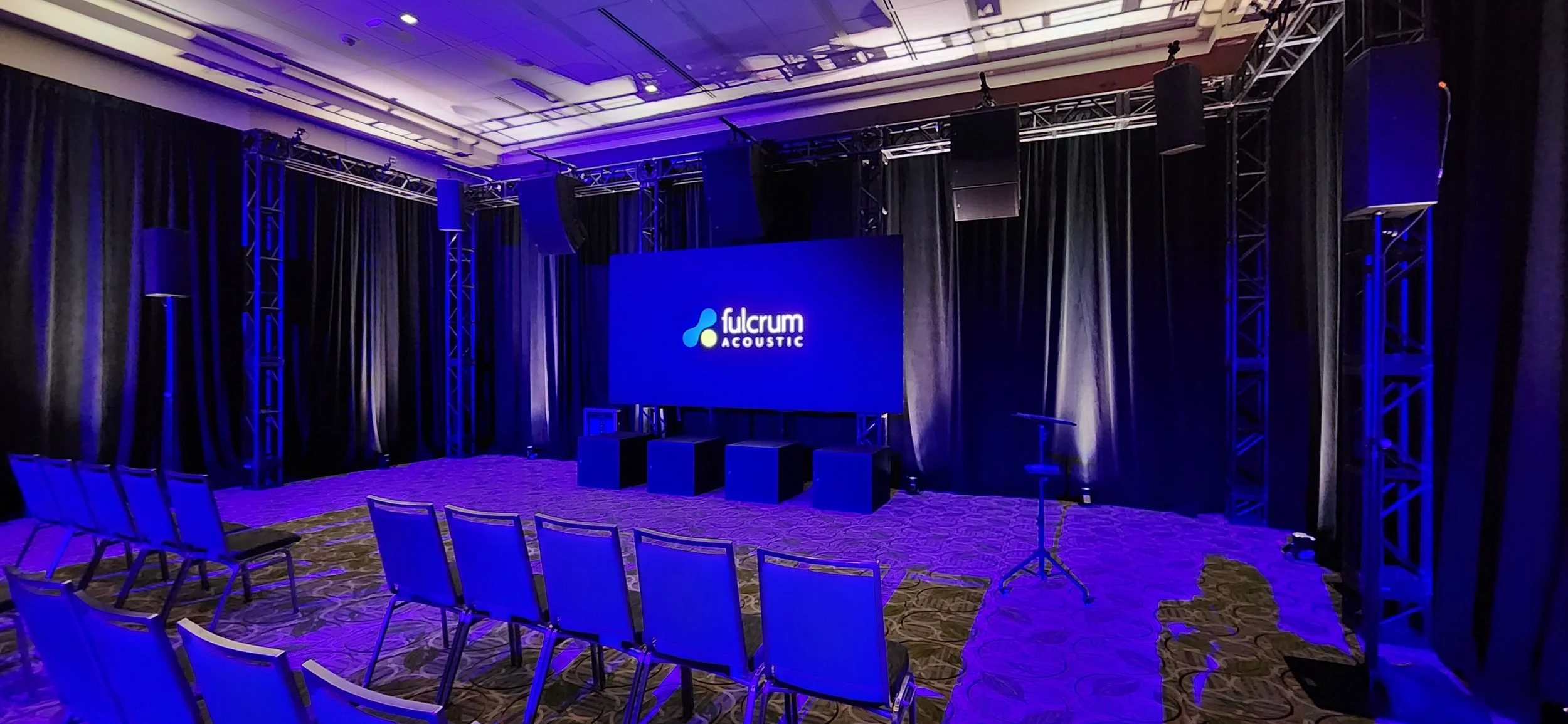 Empty conference room with black curtains, a large screen displaying 'fulcrum ACOUSTIC', and blue lighting with chairs set up facing the stage.