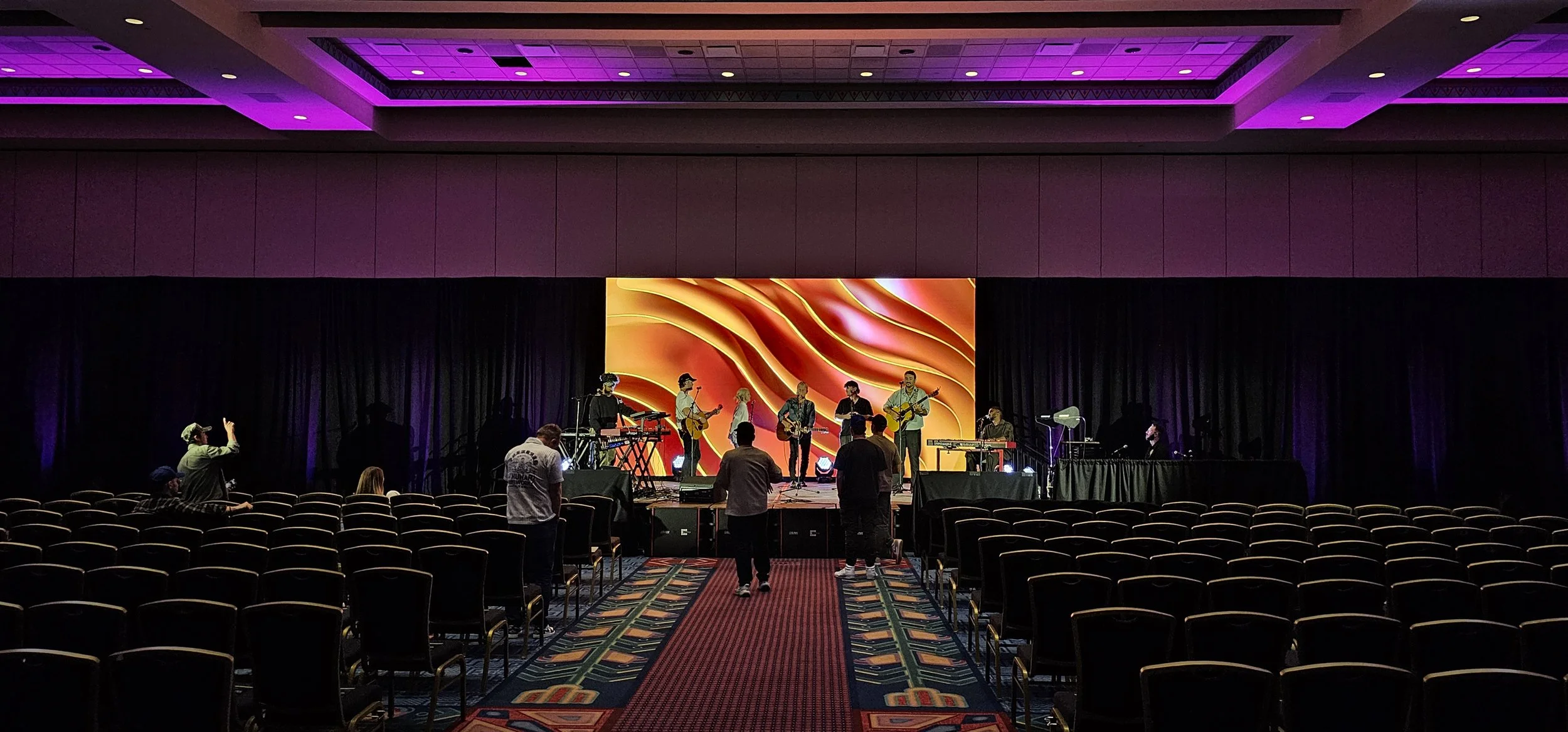 Concert stage with a band of six musicians, some playing guitars and keyboard, preparing for a performance in an indoor venue with purple lighting and a vibrant orange backdrop, empty audience seating in front.