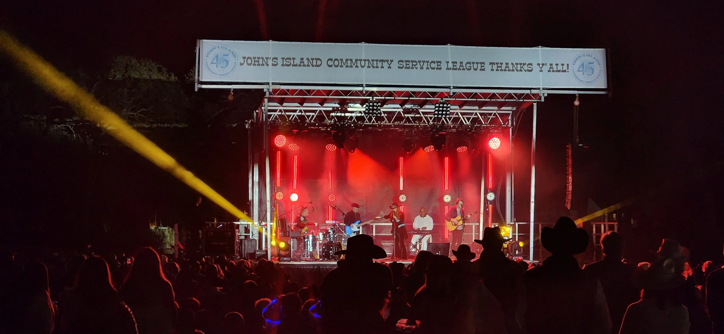 Concert on an outdoor stage lit with red and yellow lights, with a band performing and an audience watching at night.