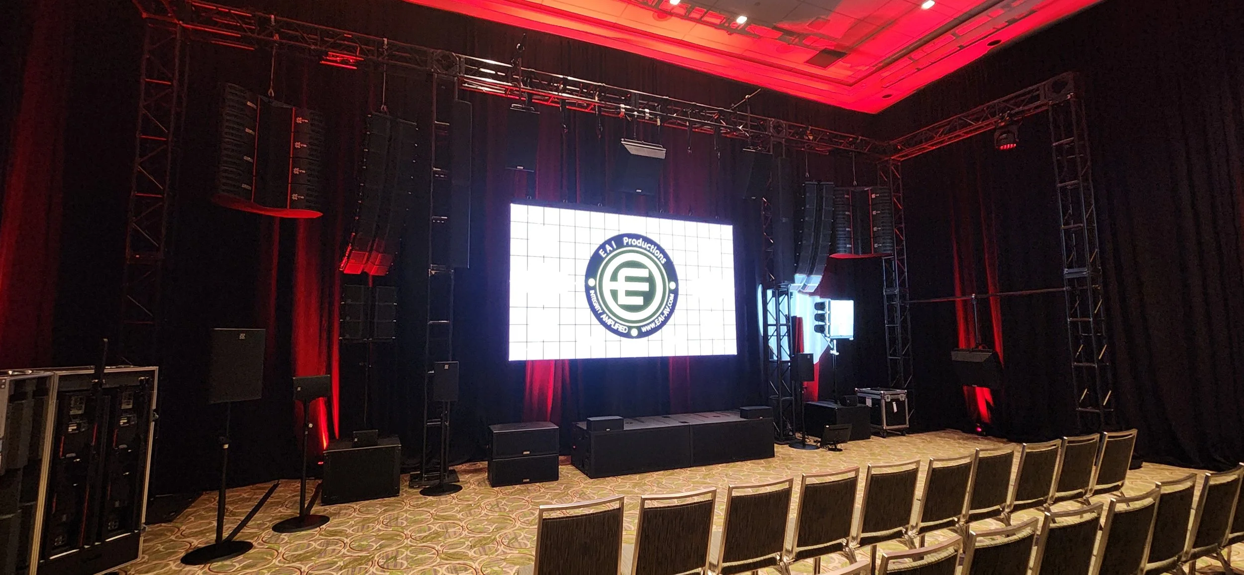 An empty conference or event hall with a large stage, black curtains, and a big digital screen displaying a logo for EAI Productions. The stage has various audio equipment, speakers, and a podium. Rows of chairs are set up facing the stage, and the l