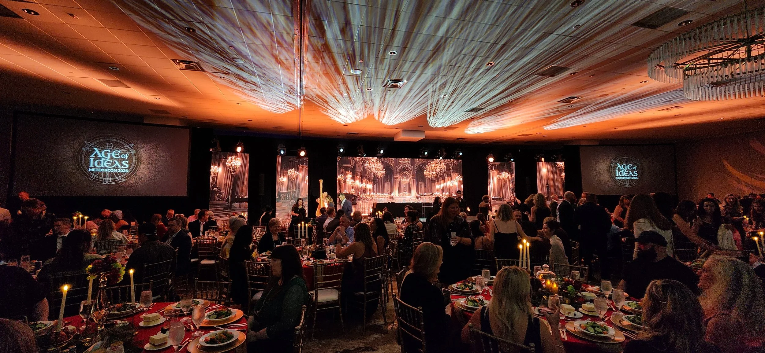 A large banquet hall with many guests seated at decorated tables, a stage with a digital backdrop featuring chandeliers, and two screens displaying the Age of Ideas logo for the Meteorcon 2025 event. The ceiling has projected patterns and lighting ef