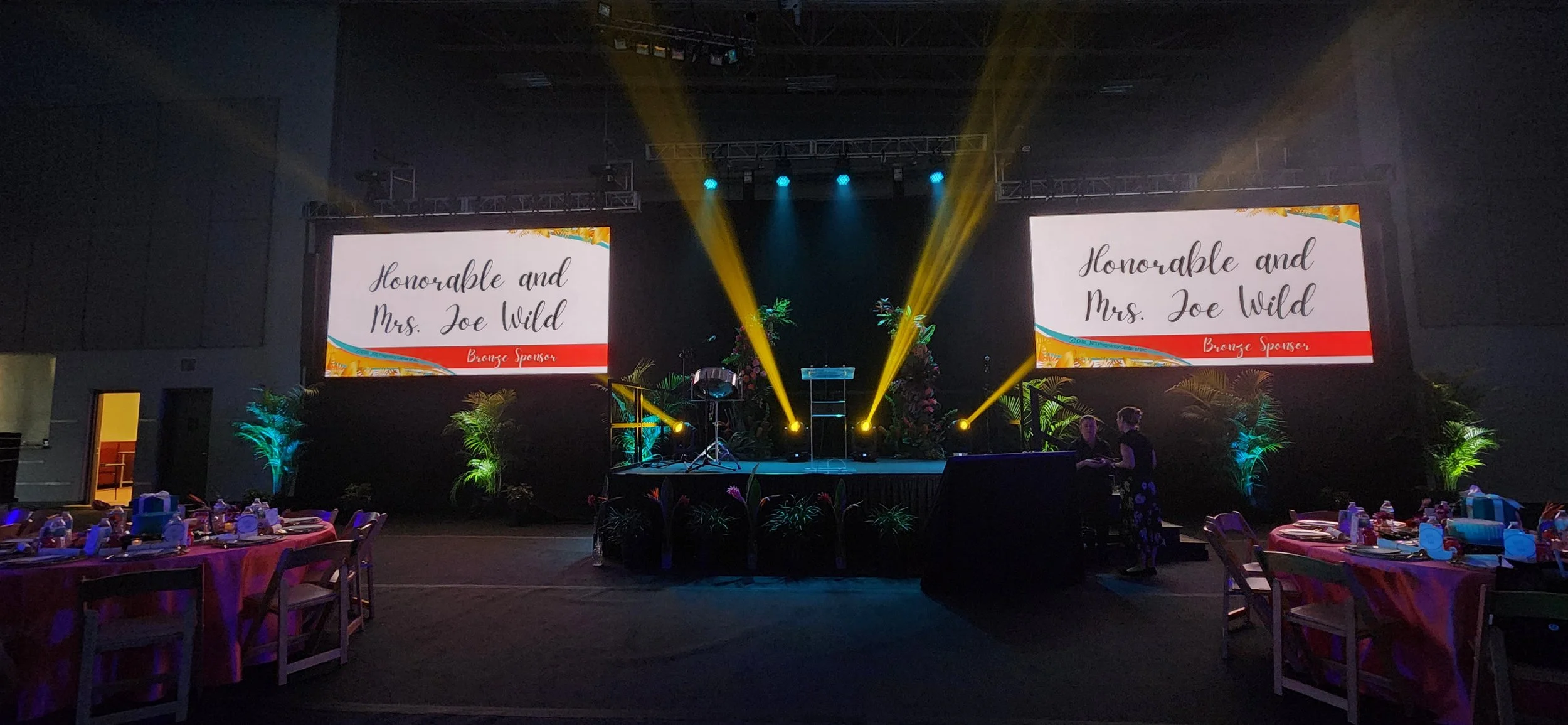 Event stage with two large screens displaying congratulations message to Honorable and Mrs. Joe Wild, bronze sponsor, decorated with plants and lighting. Tables set for a banquet are visible in the foreground.