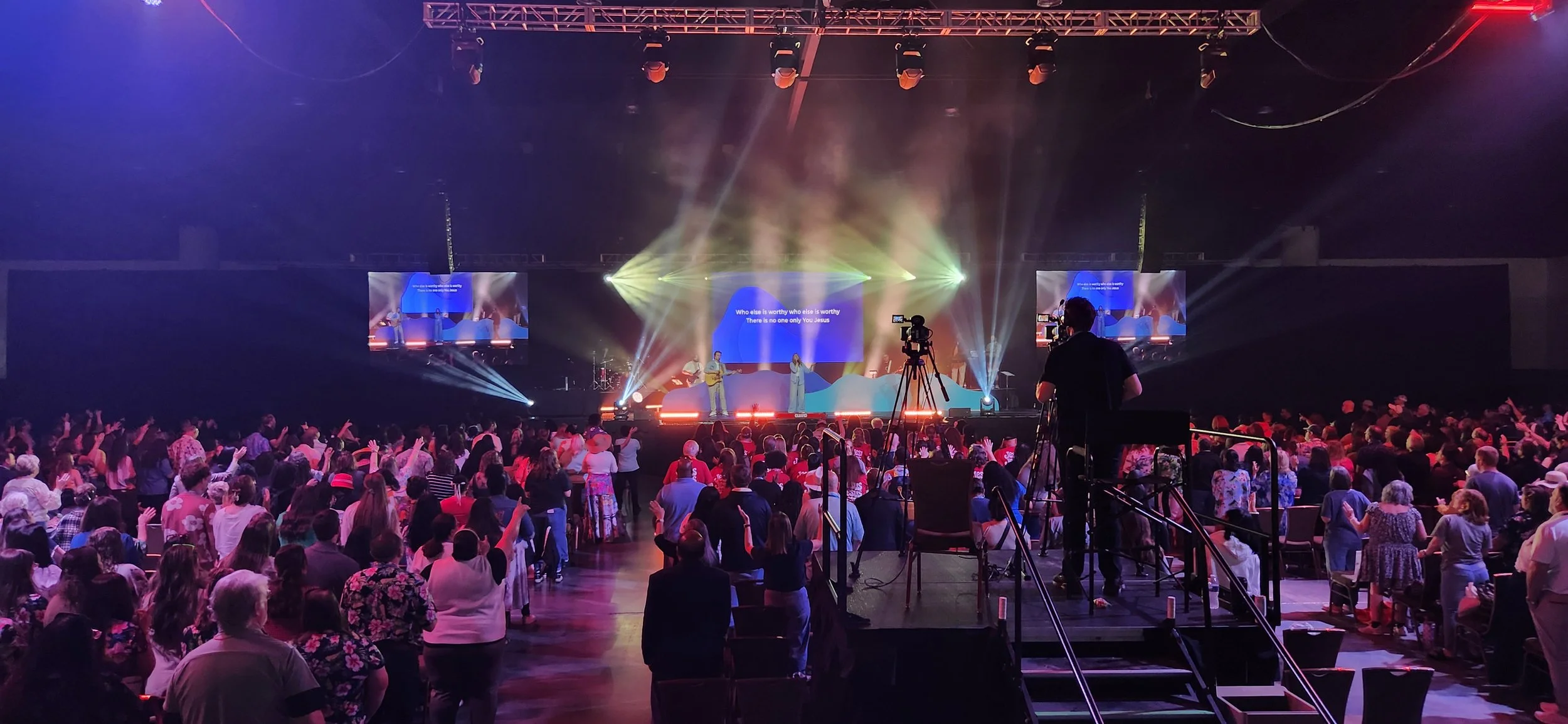 A large indoor concert venue filled with a crowd of people watching a stage performance with bright lights and large screens.