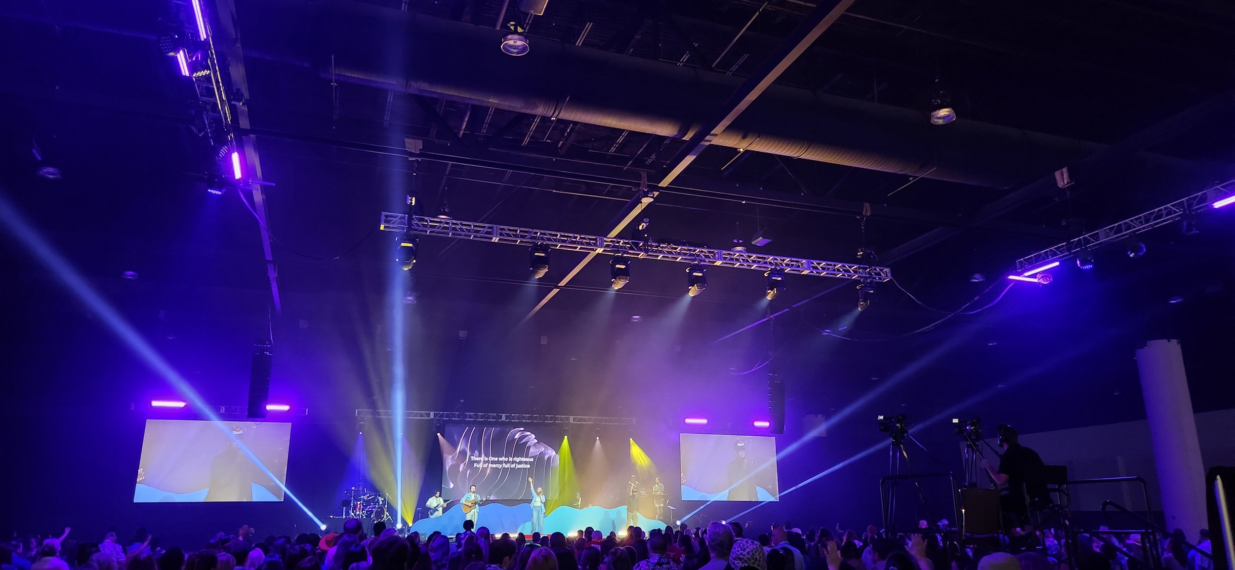 Stage with band performing, large screens displaying lyrics, colorful stage lighting, audience in foreground, event in an indoor venue.