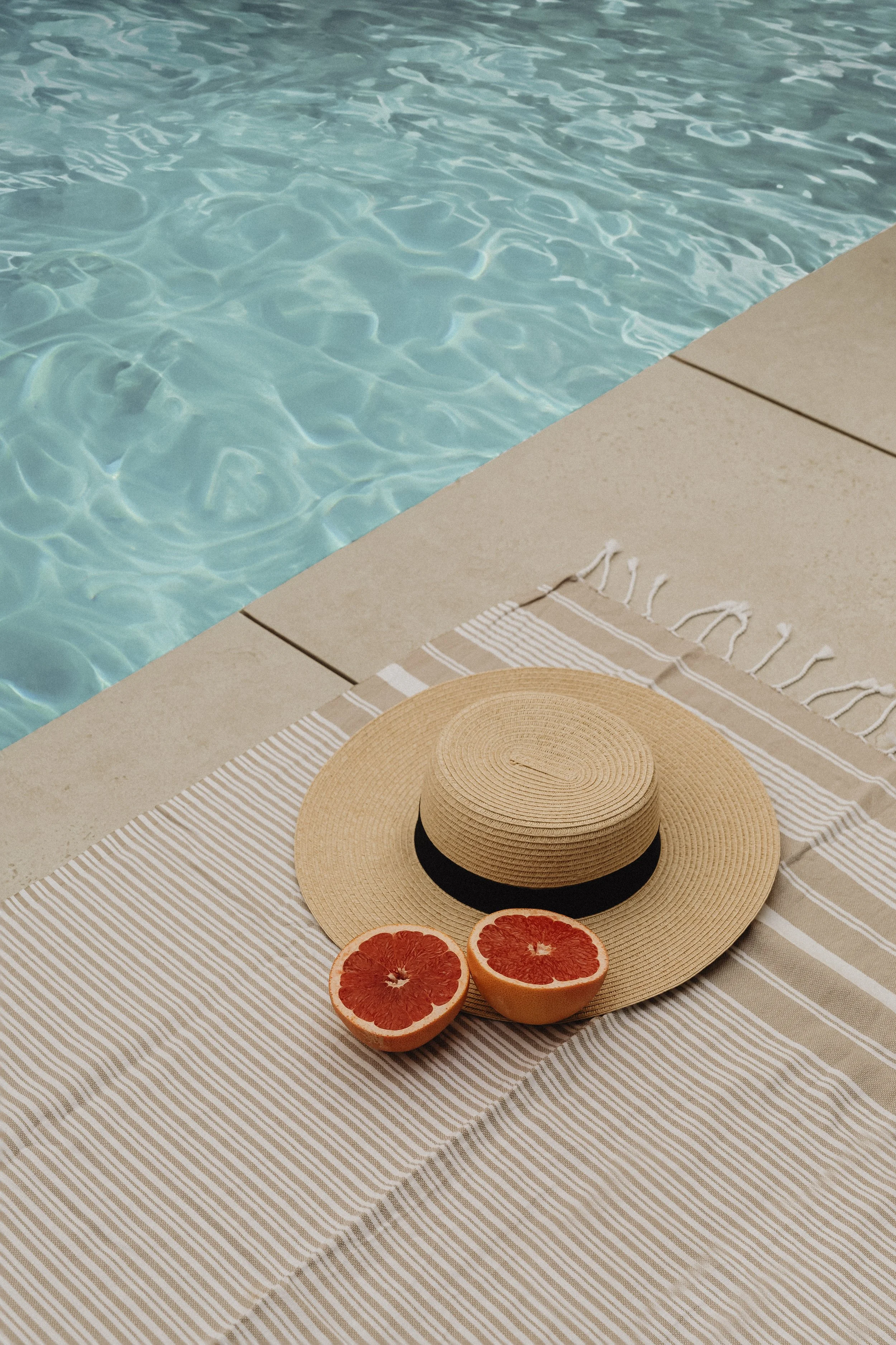 A beige straw sun hat with a black band, two halves of a grapefruit, and a beige and white striped towel are arranged on a tan poolside surface next to a swimming pool.