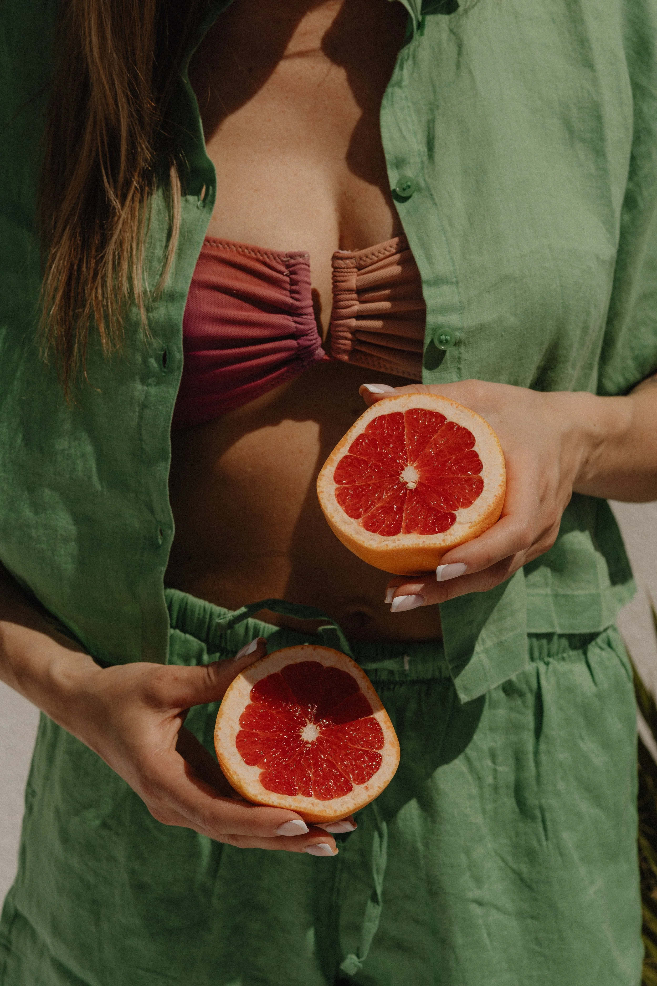 Person in green clothing holding two halved grapefruits, with a pinkish bra visible underneath an open green shirt.