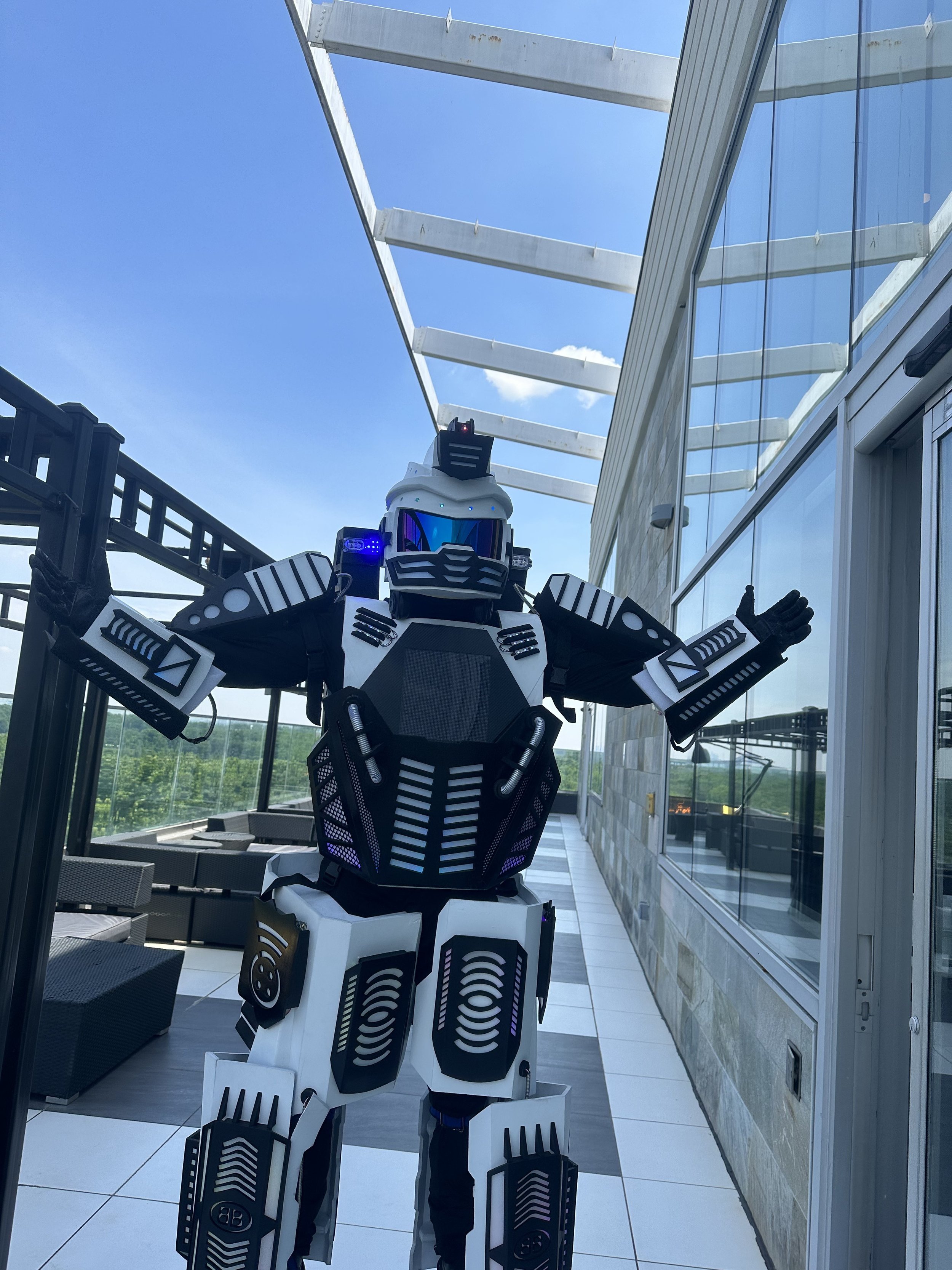 Person in a futuristic robot costume on a rooftop with glass walls and blue sky in the background.