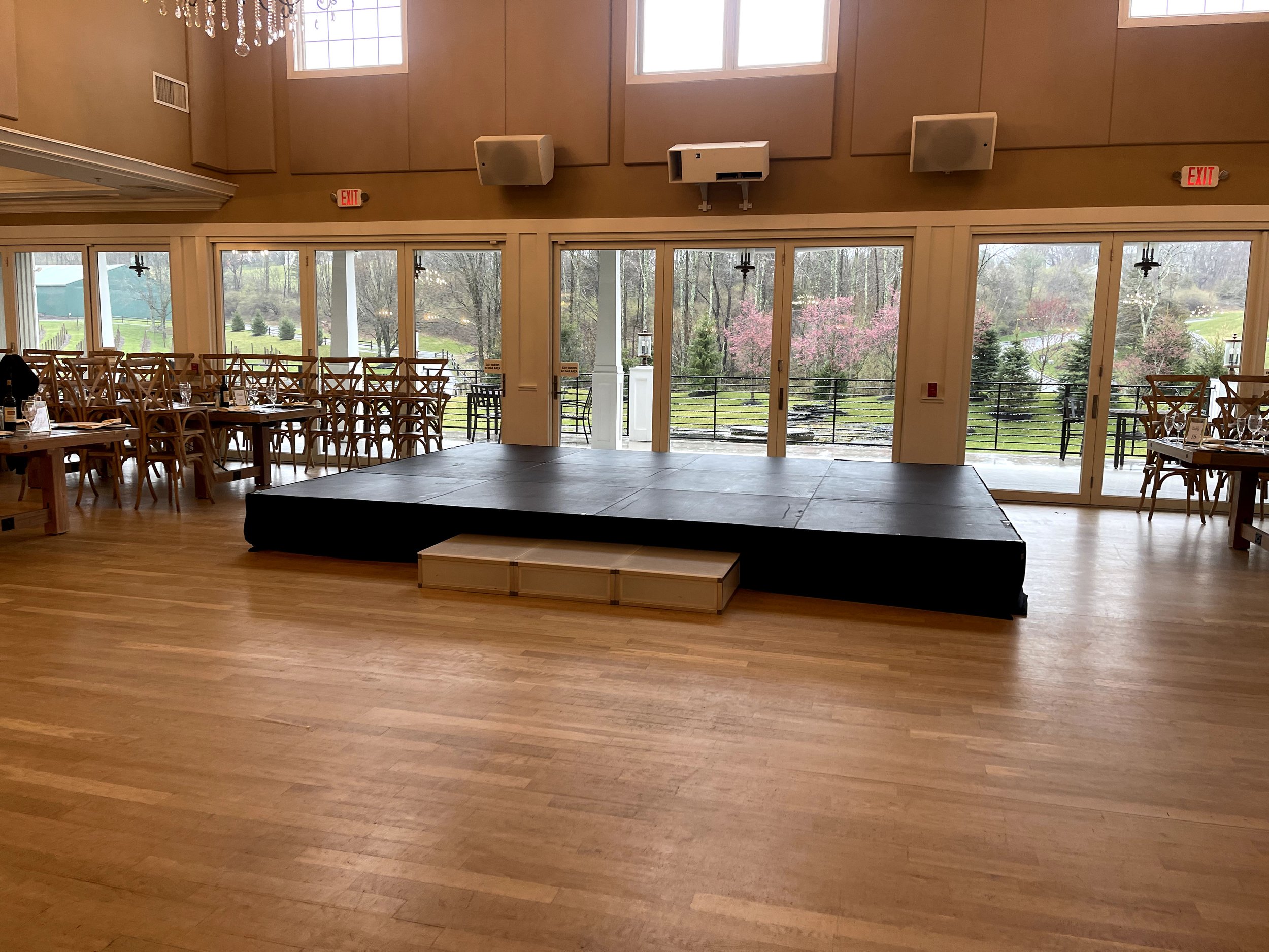 Empty event venue with a small stage, wooden chairs and tables stacked, large windows showing an outdoor view of trees and greenery.