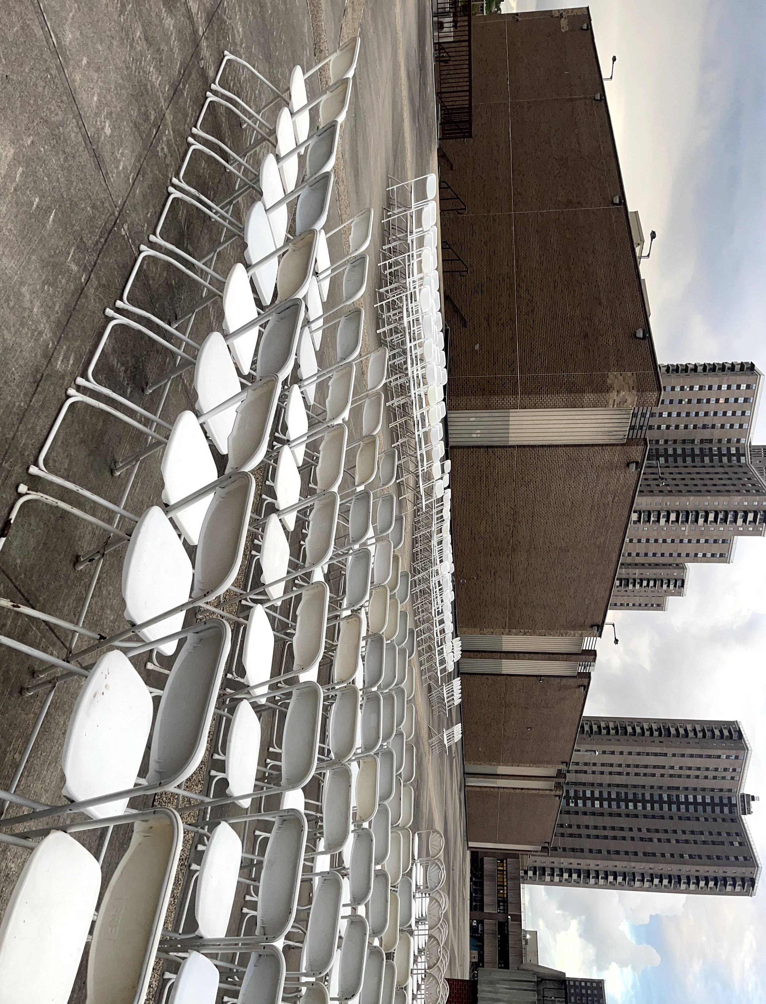 Rows of empty white folding chairs on a concrete surface with buildings in the background.