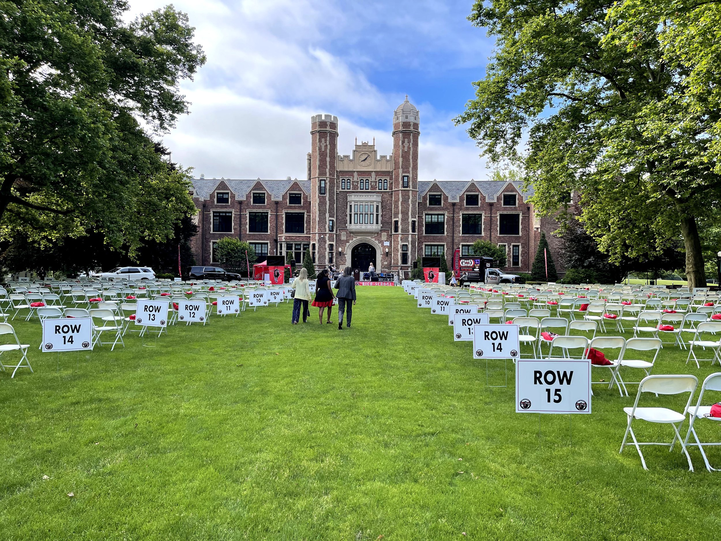 Outdoor event setup with rows of white folding chairs on a lawn, labeled by row numbers. A large brick building with towers is in the background, suggesting a formal setting like a graduation or ceremony.