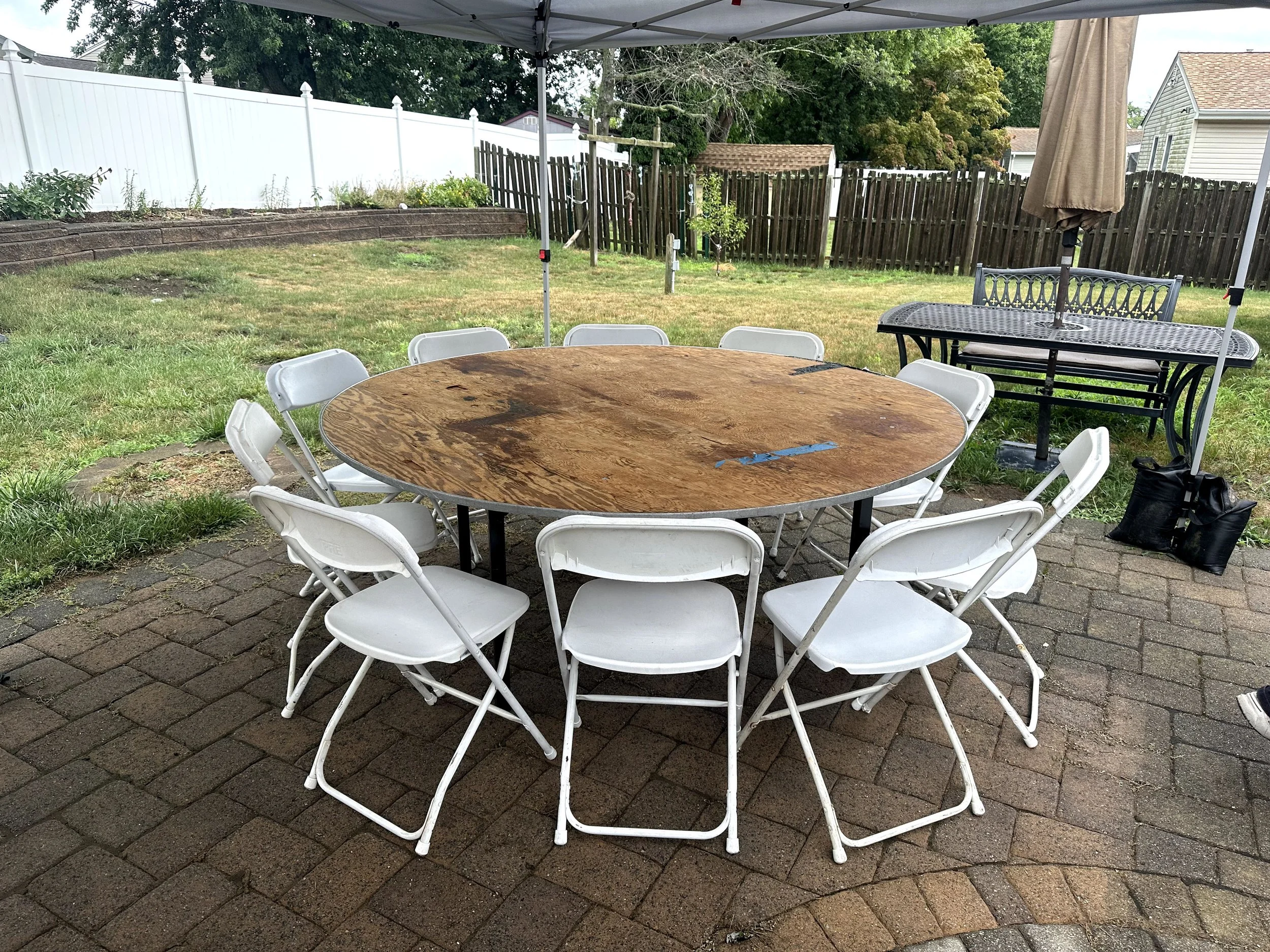 Round wooden table with white folding chairs arranged around it on a brick patio in a backyard, with a canopy overhead.