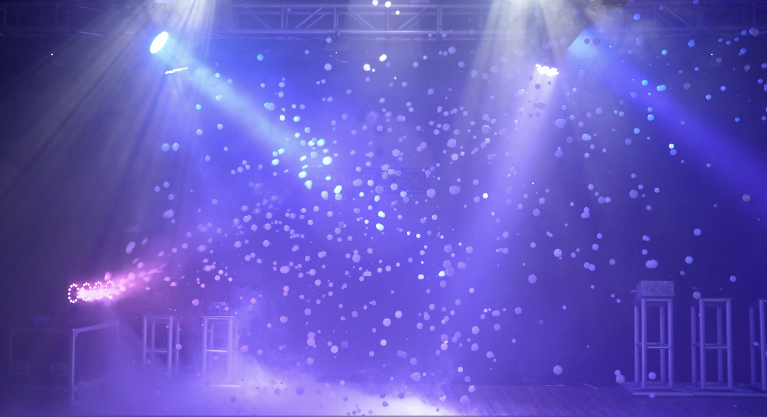 Stage with purple lighting and fog effect, white bubbles floating, and spots of light from overhead spotlights.