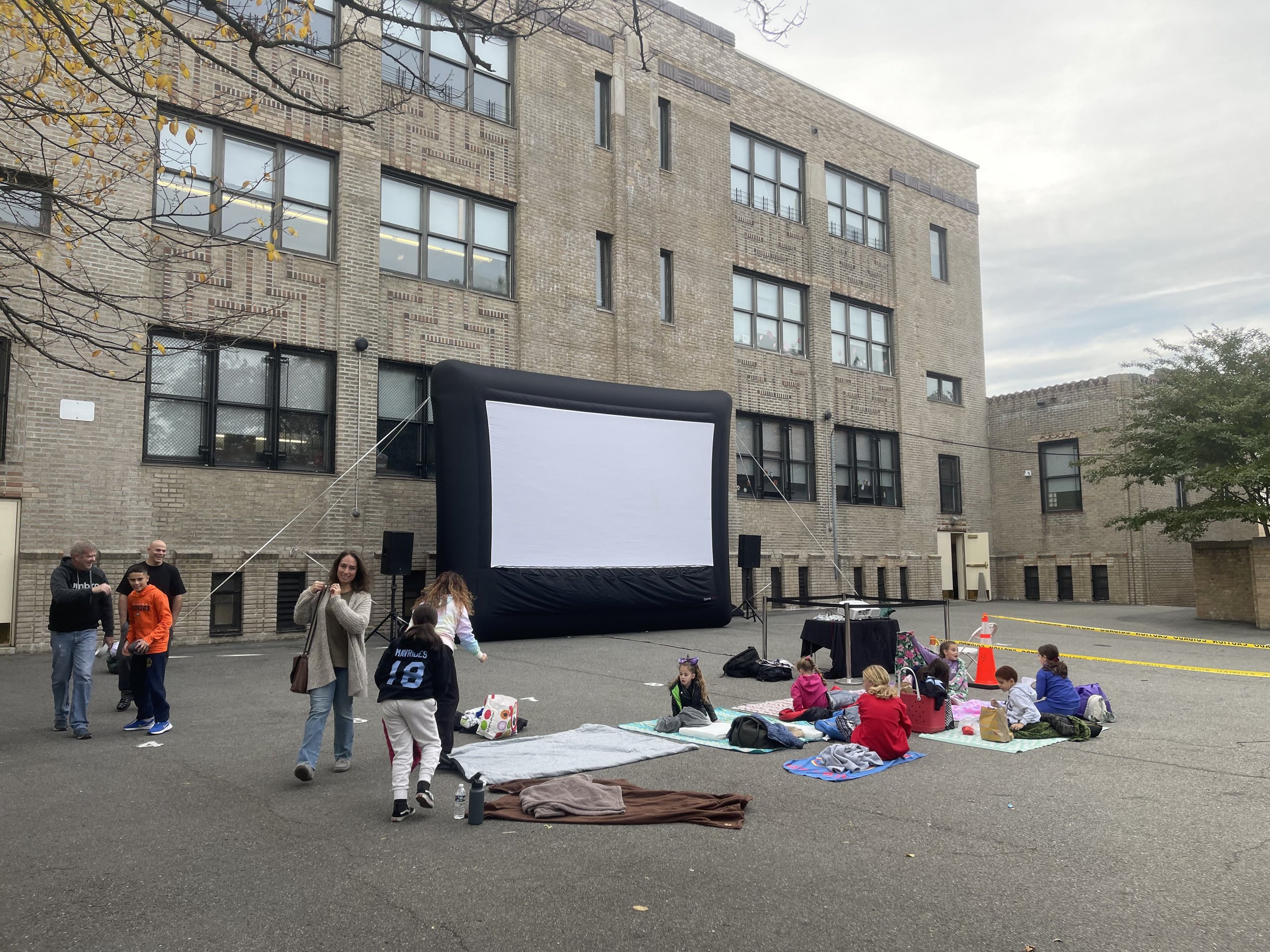 Outdoor movie setup with a large inflatable screen in a school courtyard. Children are sitting on blankets on the ground, preparing for a movie screening. Nearby, adults and kids are standing and walking. The background features a brick school buildi