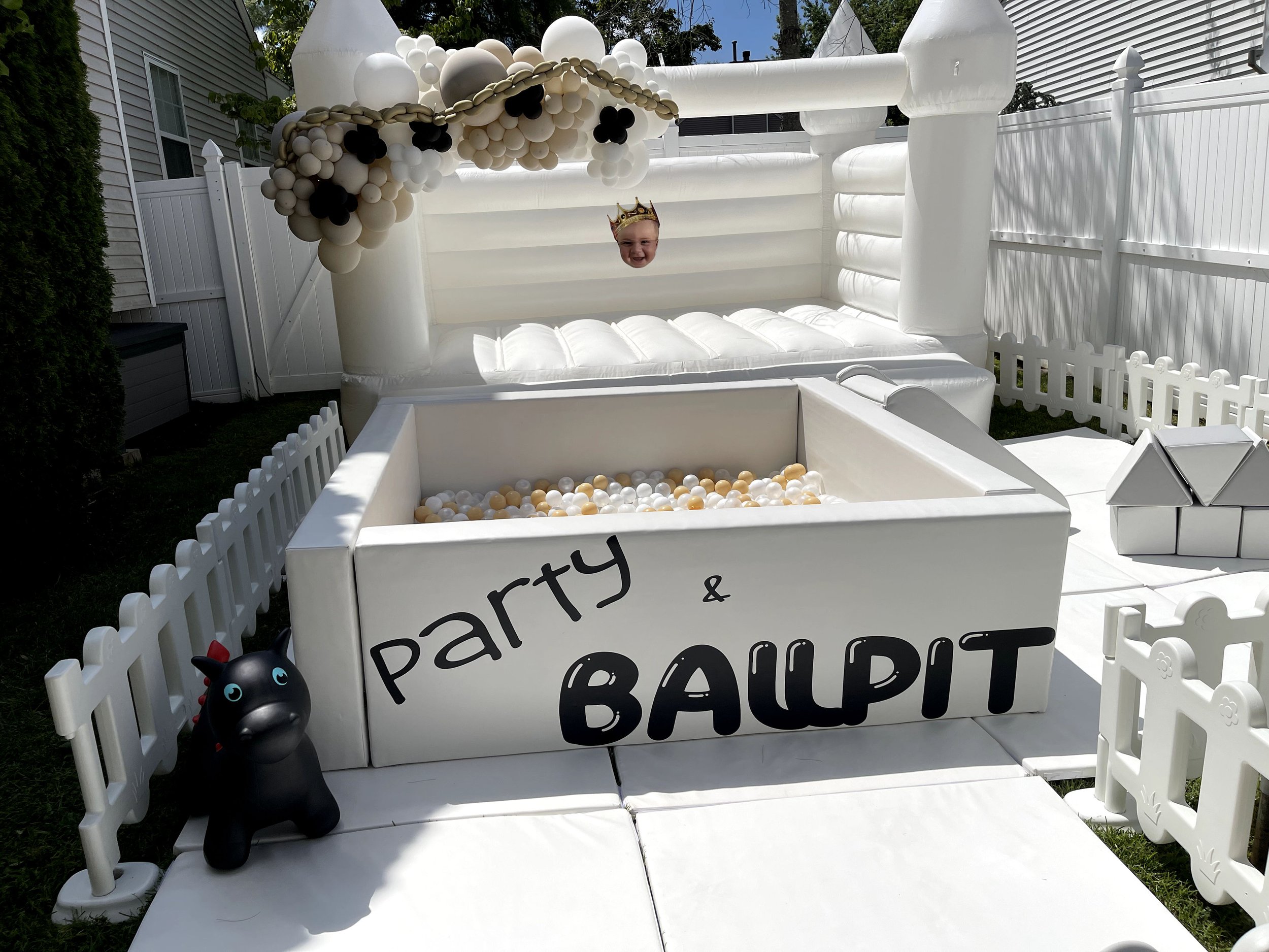 White inflatable bounce house and ball pit in a fenced yard, adorned with balloon decorations and a small black inflatable horse.
