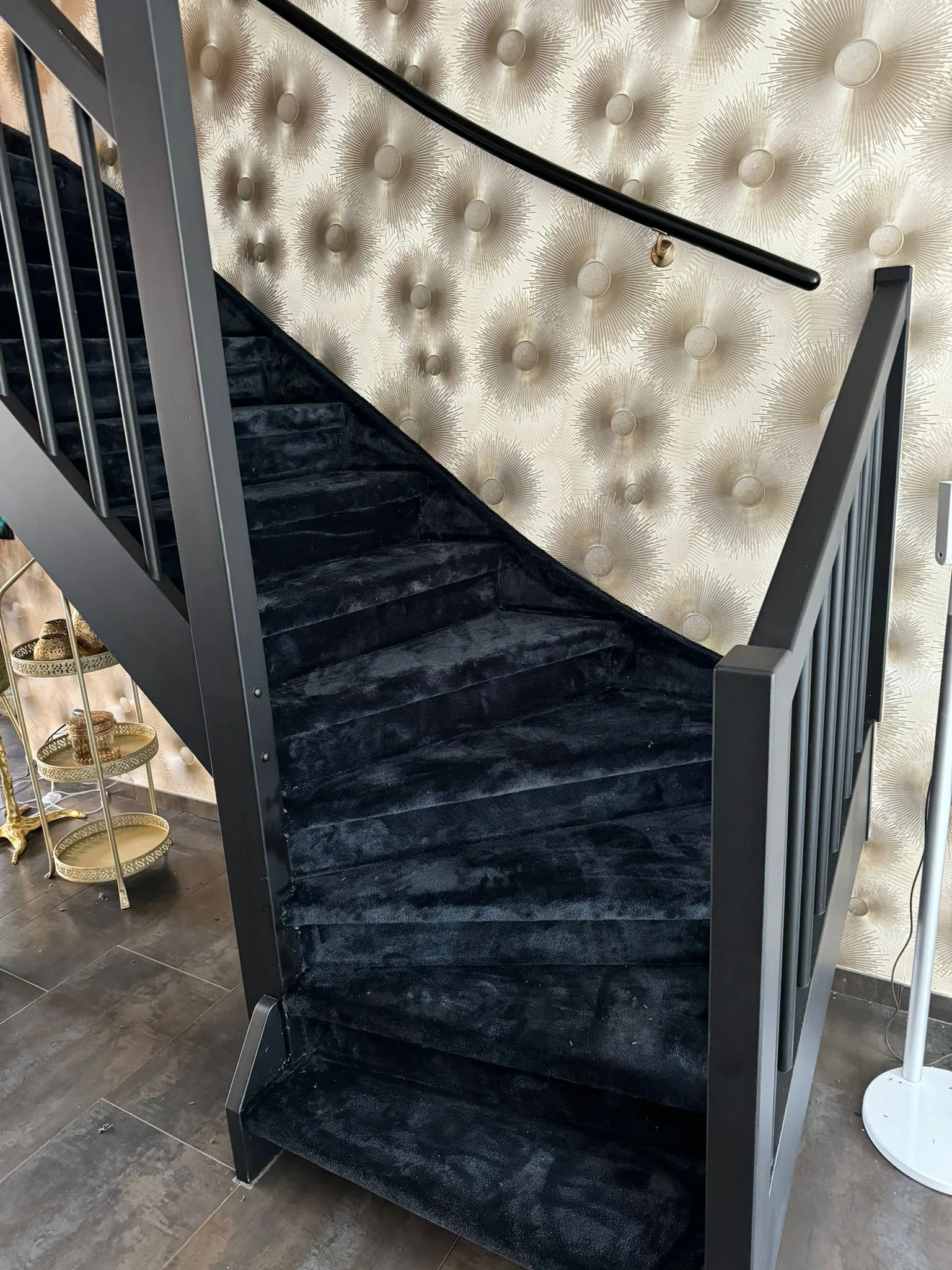 Een trap met zwarte bekleding en een metalen rand, voorgezien van een muur met een patroon in goud en wit, en een goudkleurige draperie op een dienblad.