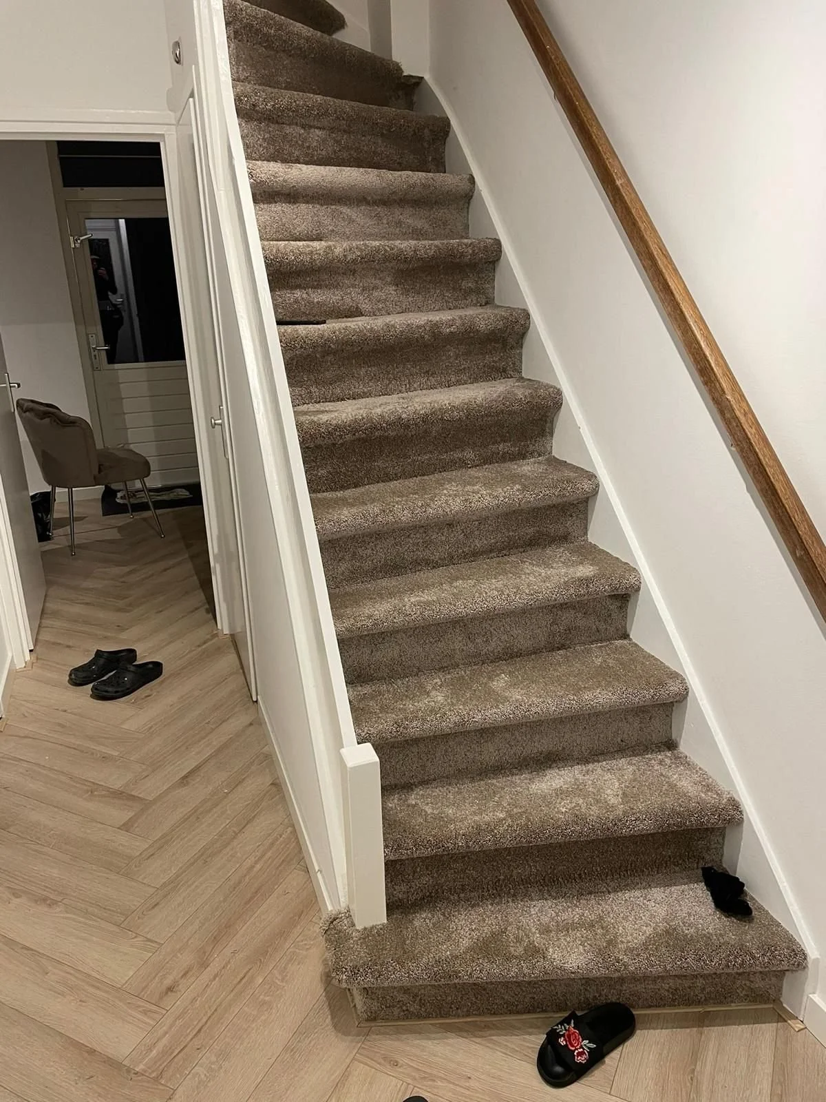 Binnenlandse trap met beige tapijt en houten handrail, naast een kamer met een stoel en schoenen op de vloer.