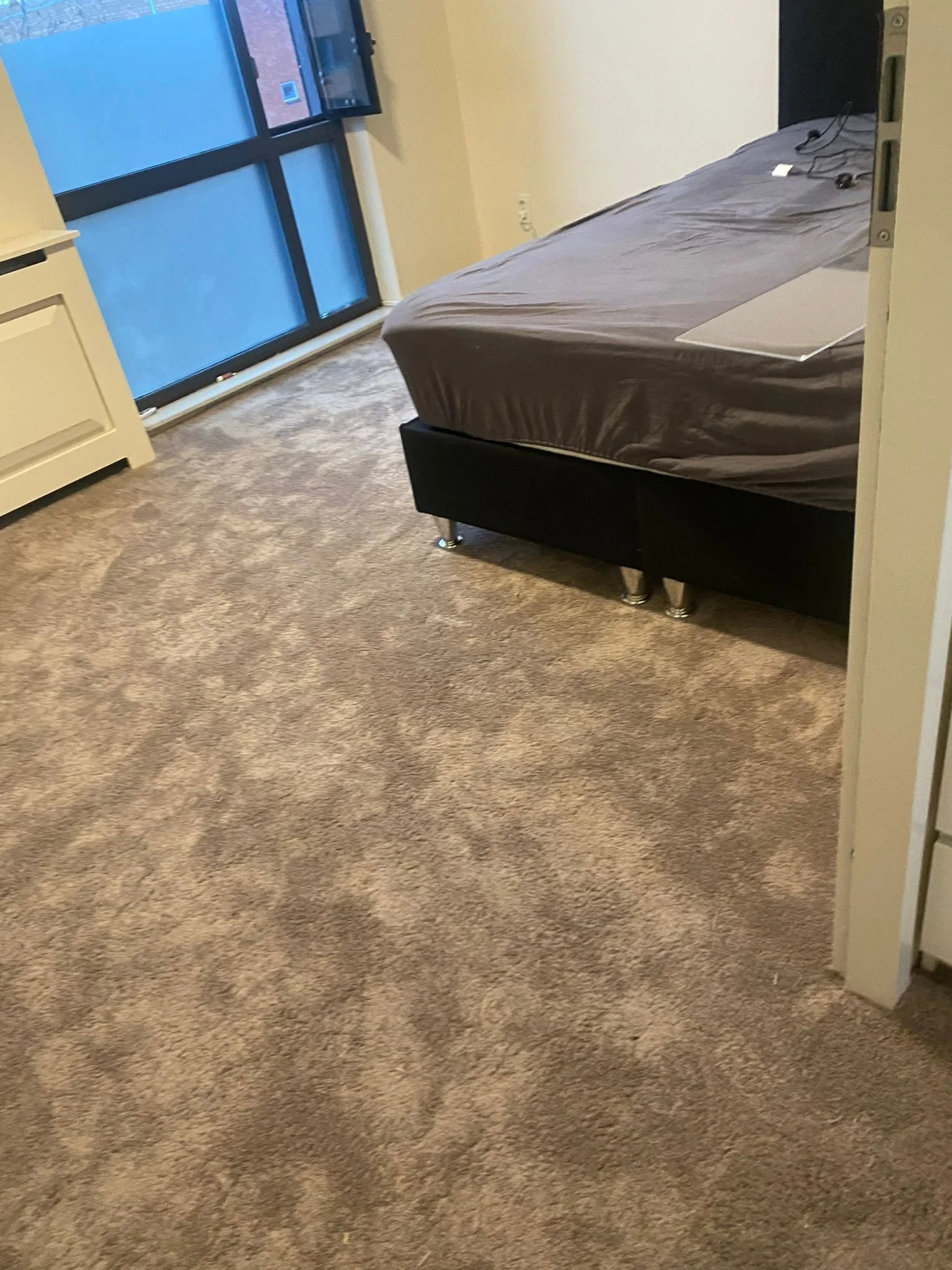 Een kamer met een bed dat bedekt is met een grijze bedspread, een vloerbedekking in bruine tinten, een raam met een luik en een televisie die uit is.