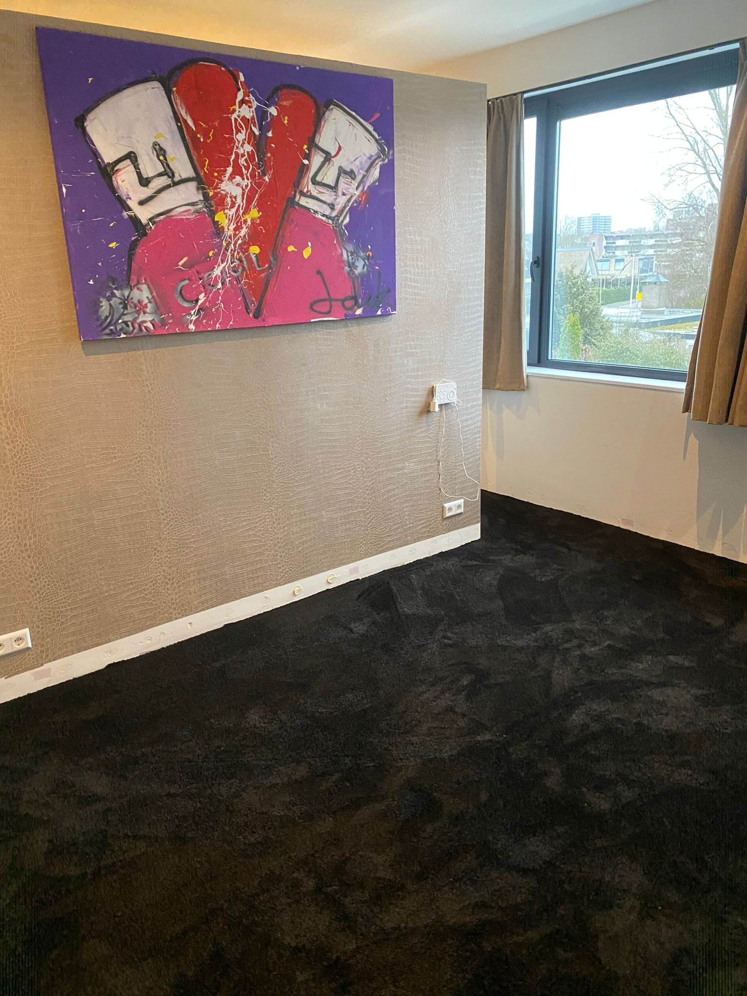 Een kamer met een muur waar een kleurrijk abstract kunstwerk hangt. Er is een groot raam met beige gordijnen dat uitzicht geeft op een stad met bomen en gebouwen. De vloer is bedekt met een zwart tapijt.