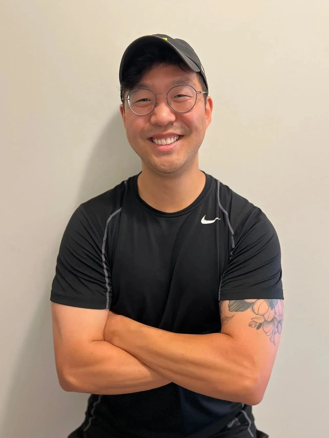 A smiling man with glasses and a cap, wearing a black athletic shirt, standing with arms crossed against a plain wall.