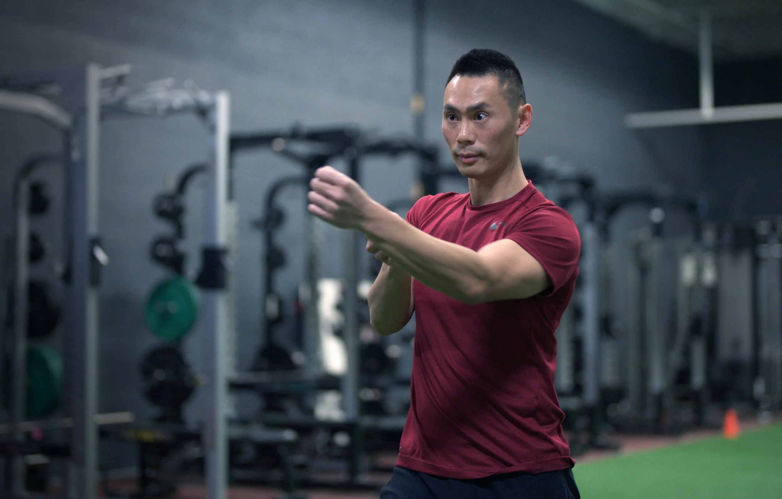 A man in a red shirt is performing a boxing move or punch in a gym. The background has gym equipment and a green artificial turf.