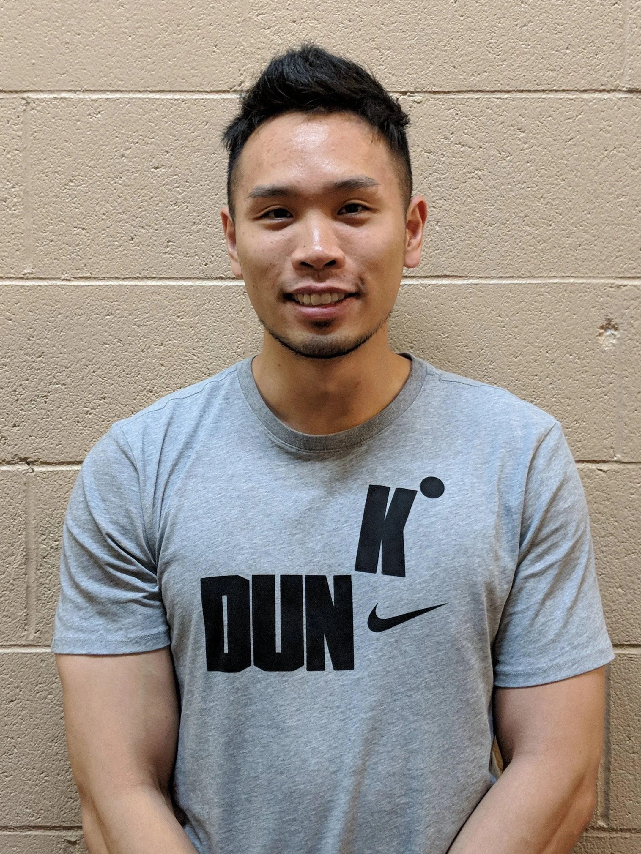 A young man with dark hair and facial hair, wearing a gray Nike t-shirt with black text and logo, standing against a beige cinder block wall, smiling at the camera.