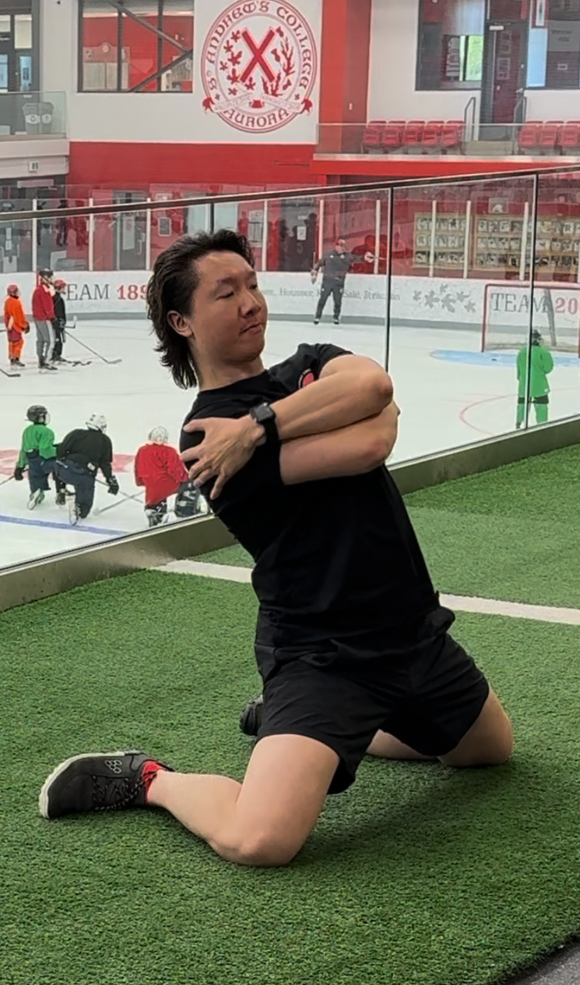 A man kneels on a patch of artificial grass in front of an ice hockey rink, hugging himself with his eyes closed. The rink's glass panels and players in hockey gear are visible behind him, along with a large logo and banners on the walls.