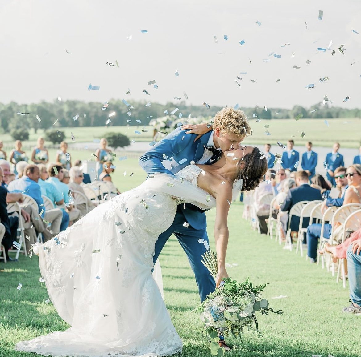 Bride and groom kissing outdoors, surrounded by confetti, with guests seated in the background at a wedding ceremony.