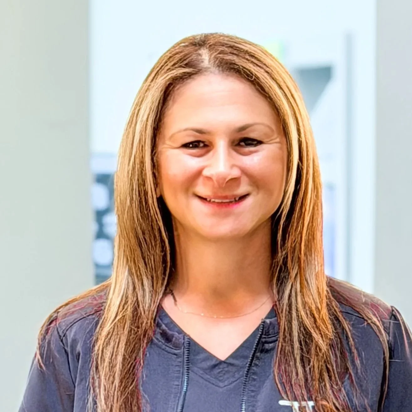 A woman with curly brown hair tied back, wearing a blue medical uniform with the logo of Turlock Dental Care, smiling and talking in an office with a computer monitor in the background displaying Minnie Mouse.