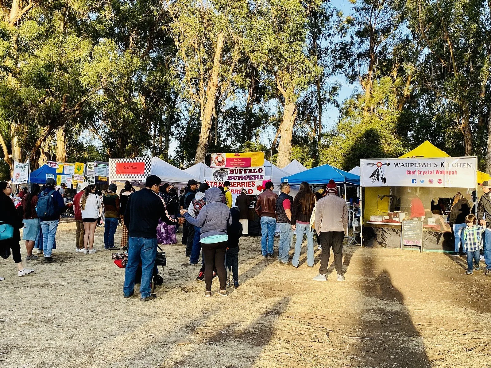 At dusk, spectators and dancers in regalia gather around the arena beneath tall eucalyptus trees, with vendor tents behind.