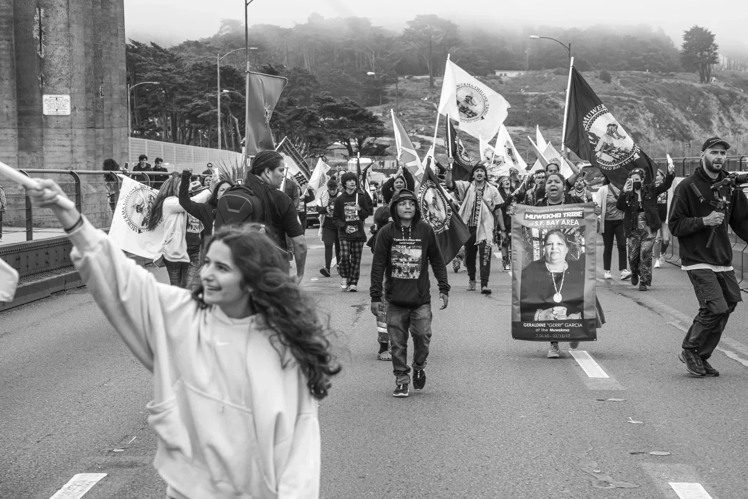 Black-and-white photo of a crowd marching down a road with flags and banners, a young woman in front raising her arm.
