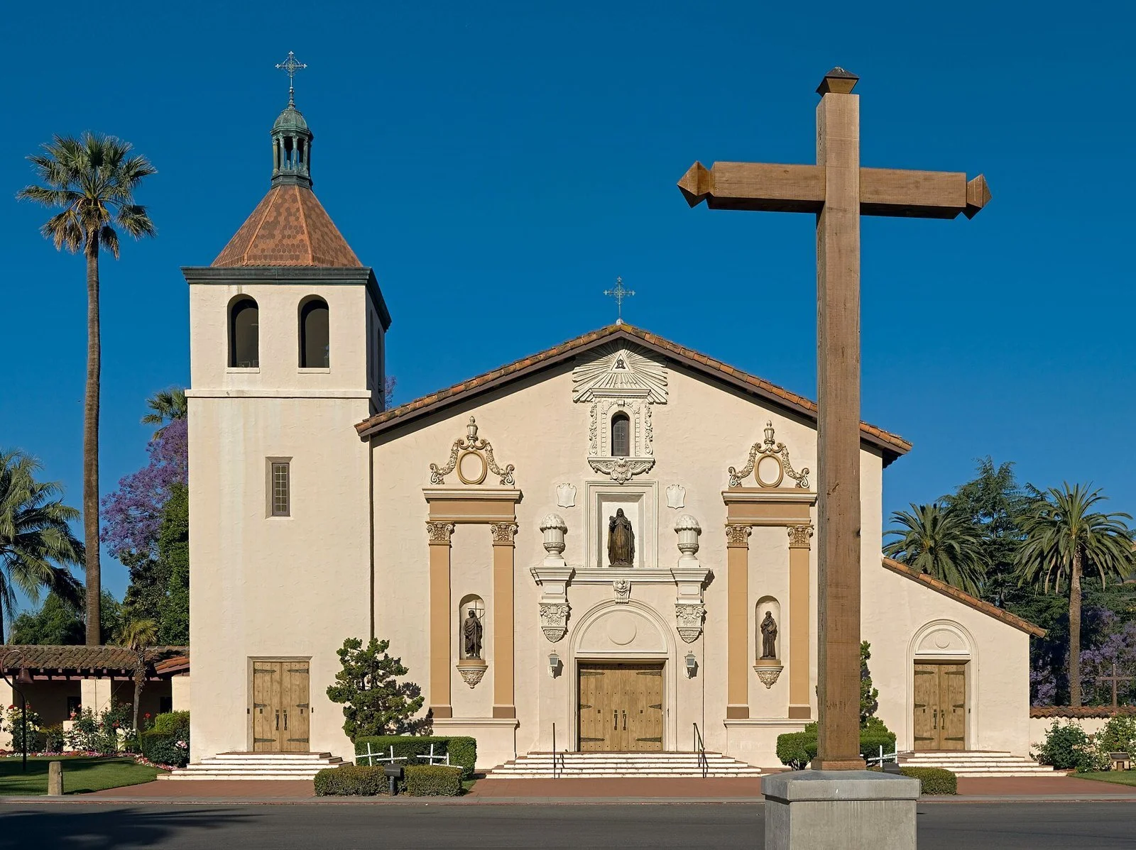 The facade of the Mission Church at Santa Clara University, with its painted saints and bell tower, behind a lawn and roses.