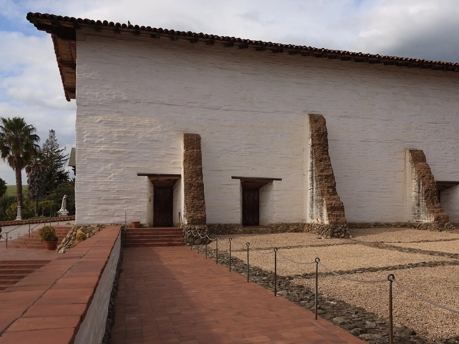 The whitewashed adobe wall and buttresses of Mission San José in Fremont, beside a brick walkway, on a clear spring day.