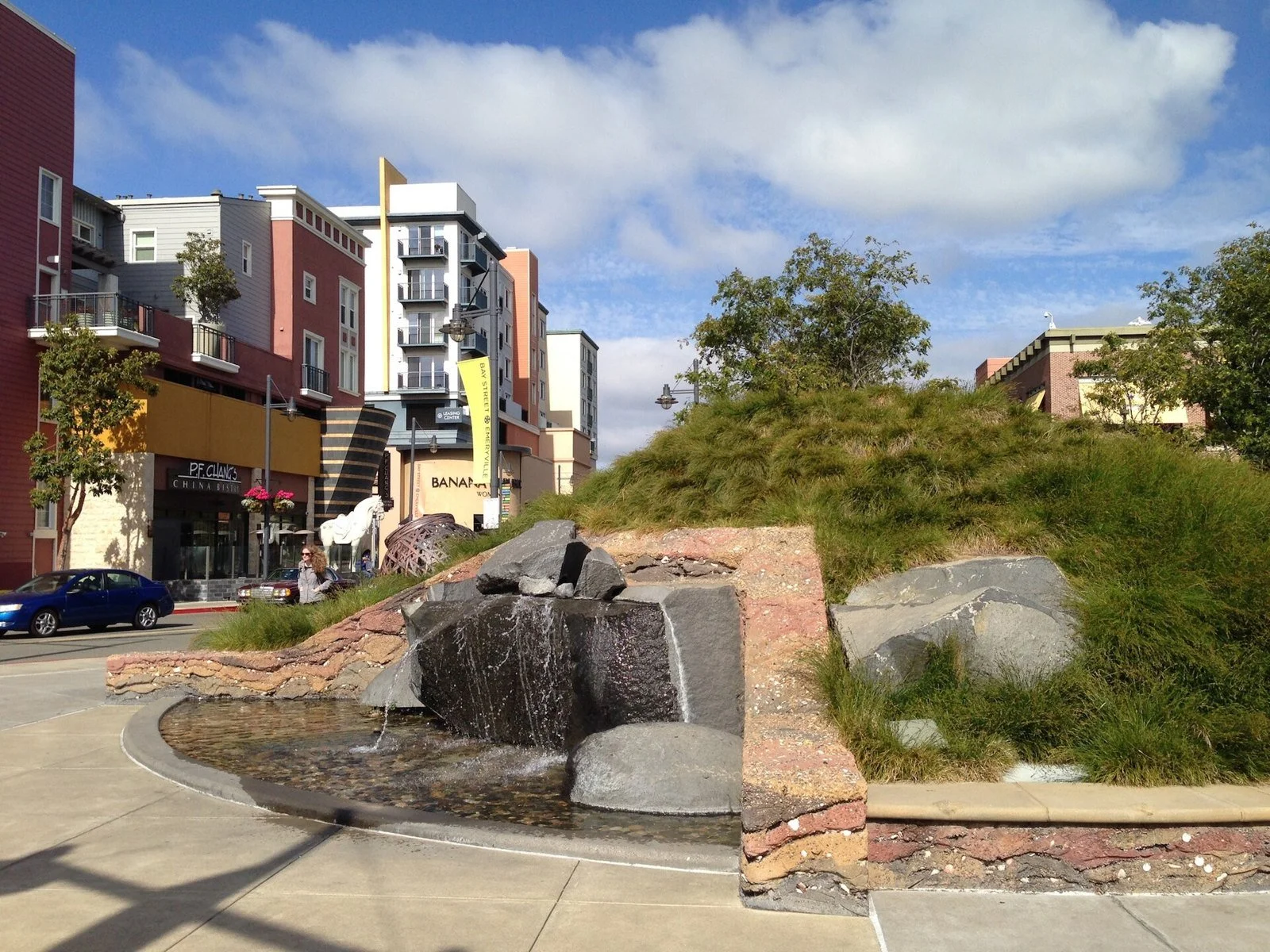 A memorial sculpture and plantings at the site of the Emeryville Shellmound, surrounded by modern buildings.