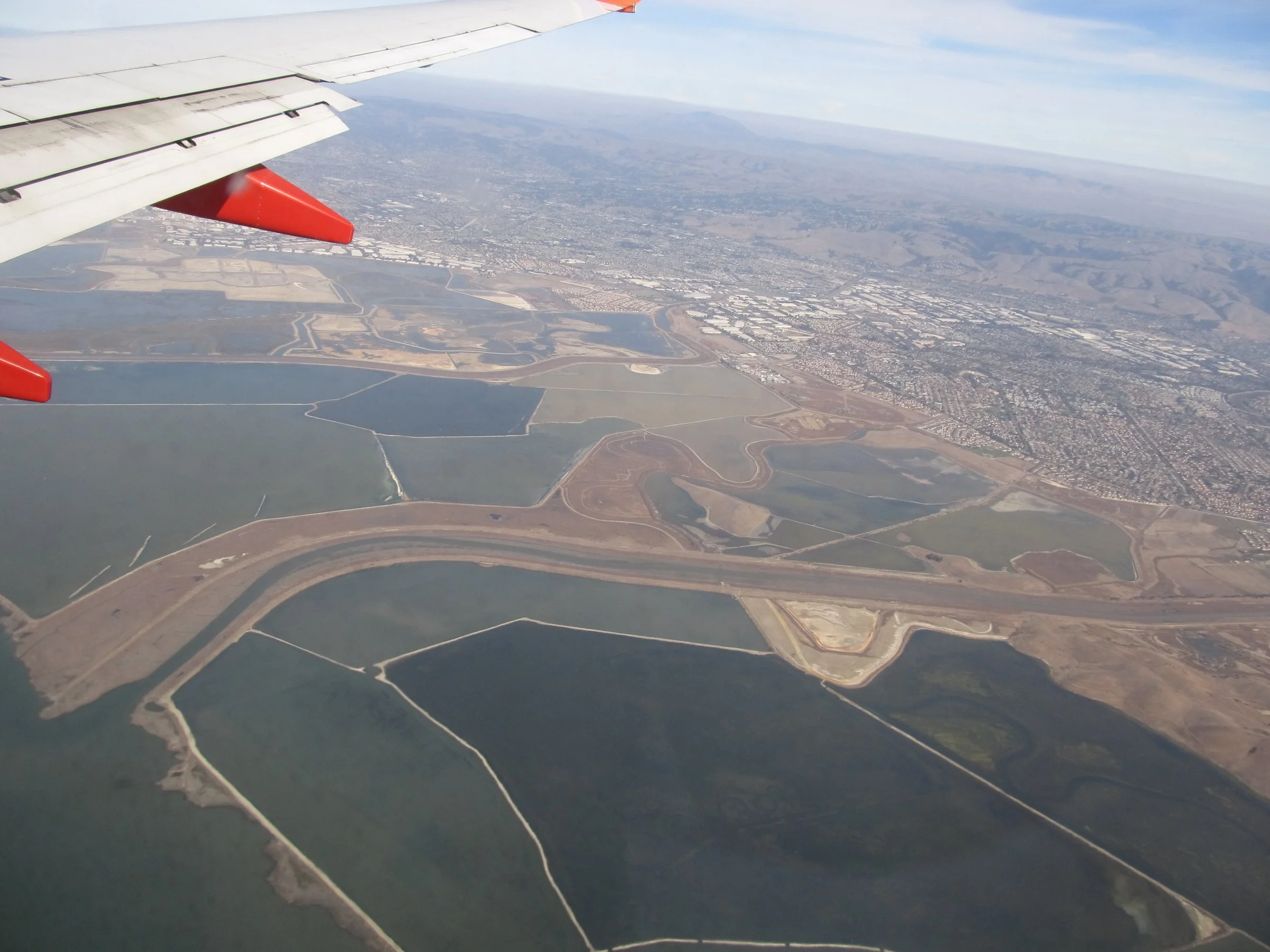 An aerial view of Eden Landing's former salt ponds, laid out in dark rectangles along a winding levee, with an airplane wing at the top of the frame.