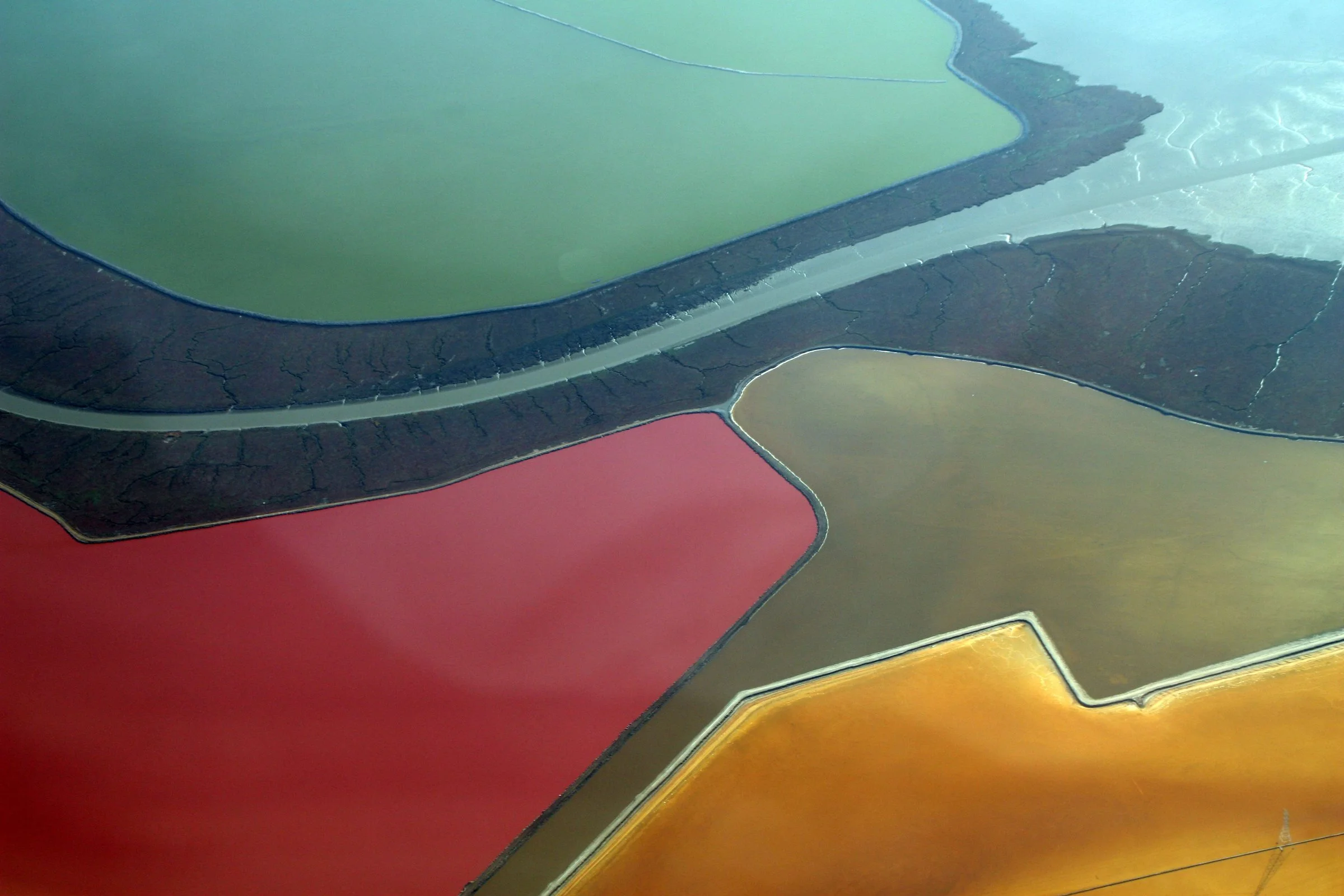 An aerial view of South Bay salt evaporation ponds in vivid red, orange, ochre and green, separated by dark levees and channels.