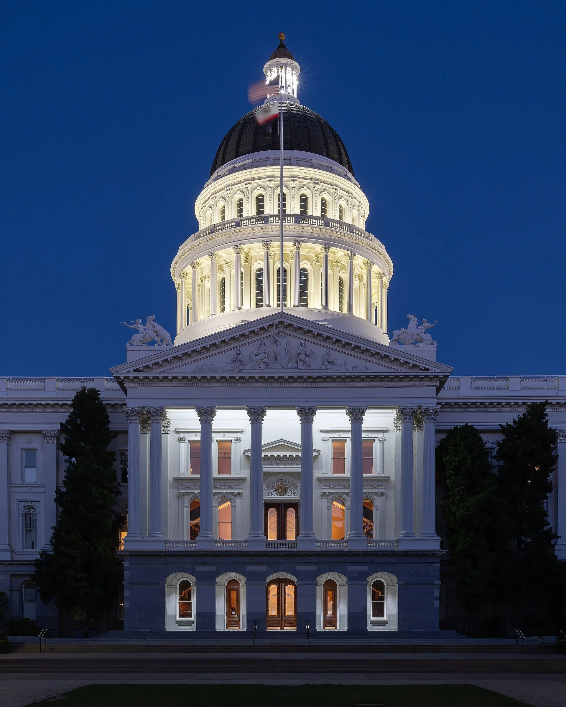 The California State Capitol at blue hour, its lit dome and white columned facade glowing against a deep blue evening sky, with dark trees on either side.