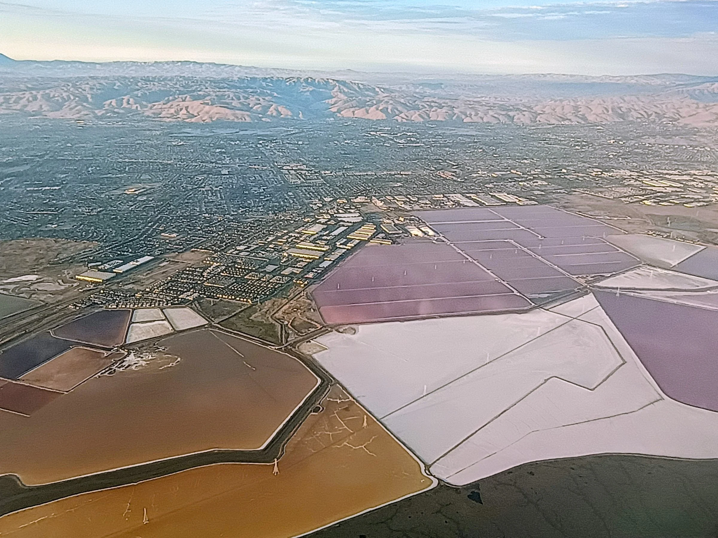 Pink, white, ochre and violet salt ponds spread across the South Bay plain below a hazy line of hills, seen from an airplane.